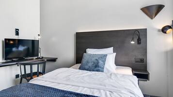 A modern hotel room with a bed, a desk with a television, a wall-mounted lamp, and a power outlet.