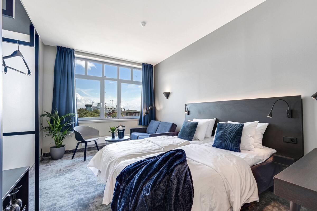 A modern hotel room with a large bed, a desk, a chair, and a view of the city through the window.