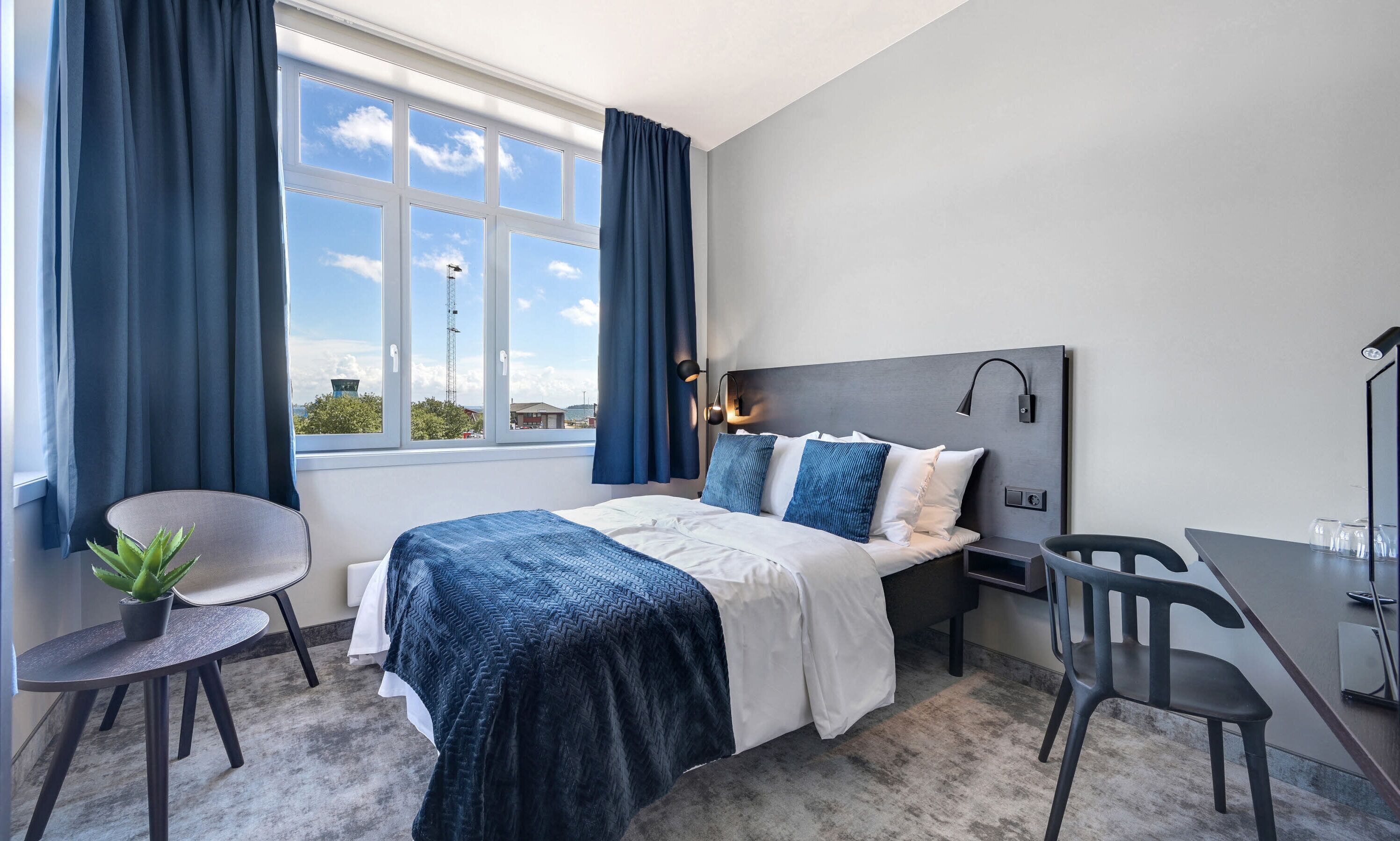 Una habitación de hotel moderna con una cama grande, un escritorio y una silla. Hay una ventana con cortinas, una planta y vistas al exterior.