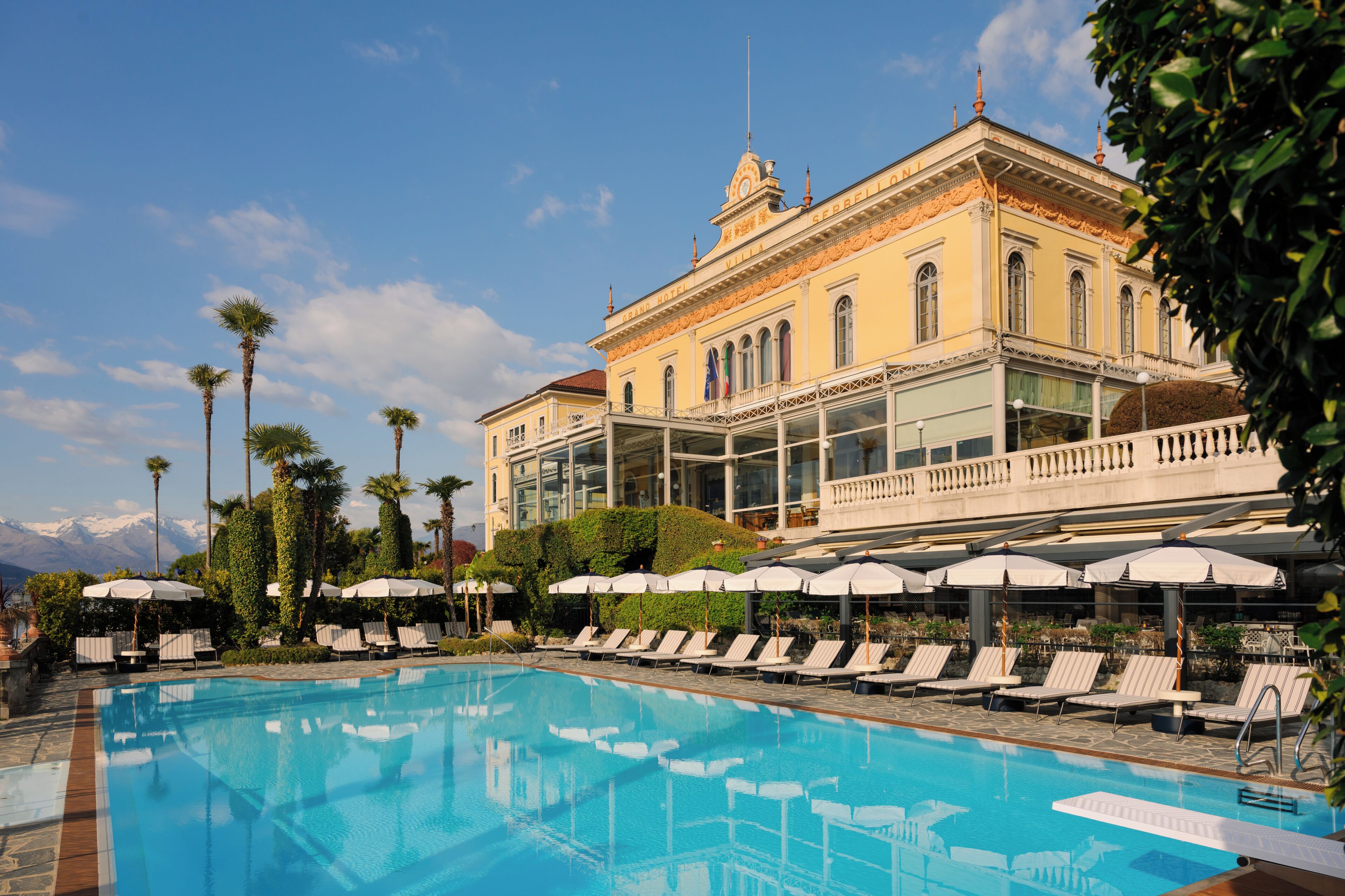 A pool with lounge chairs and umbrellas, a building with classical architecture, and a clear sky.