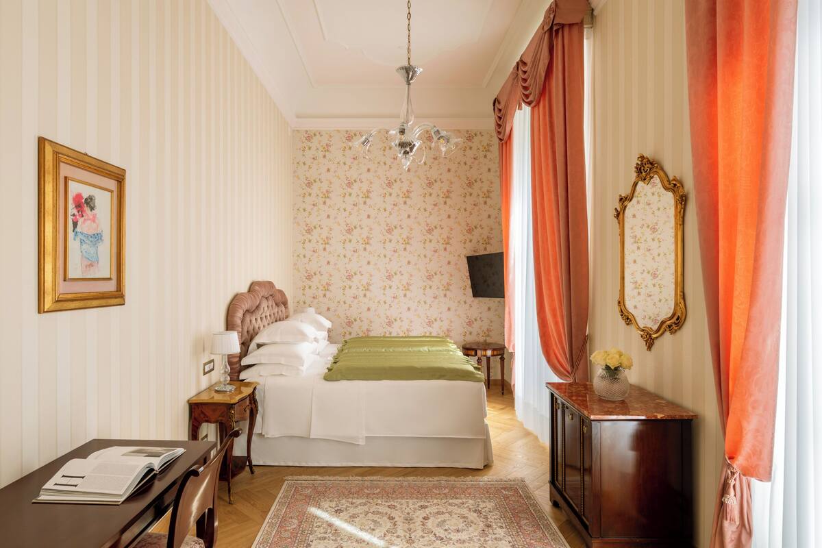 Une chambre d’hôtel avec un lit, un bureau sur lequel se trouve un livre, une chaise, une commode, un miroir et un lustre.