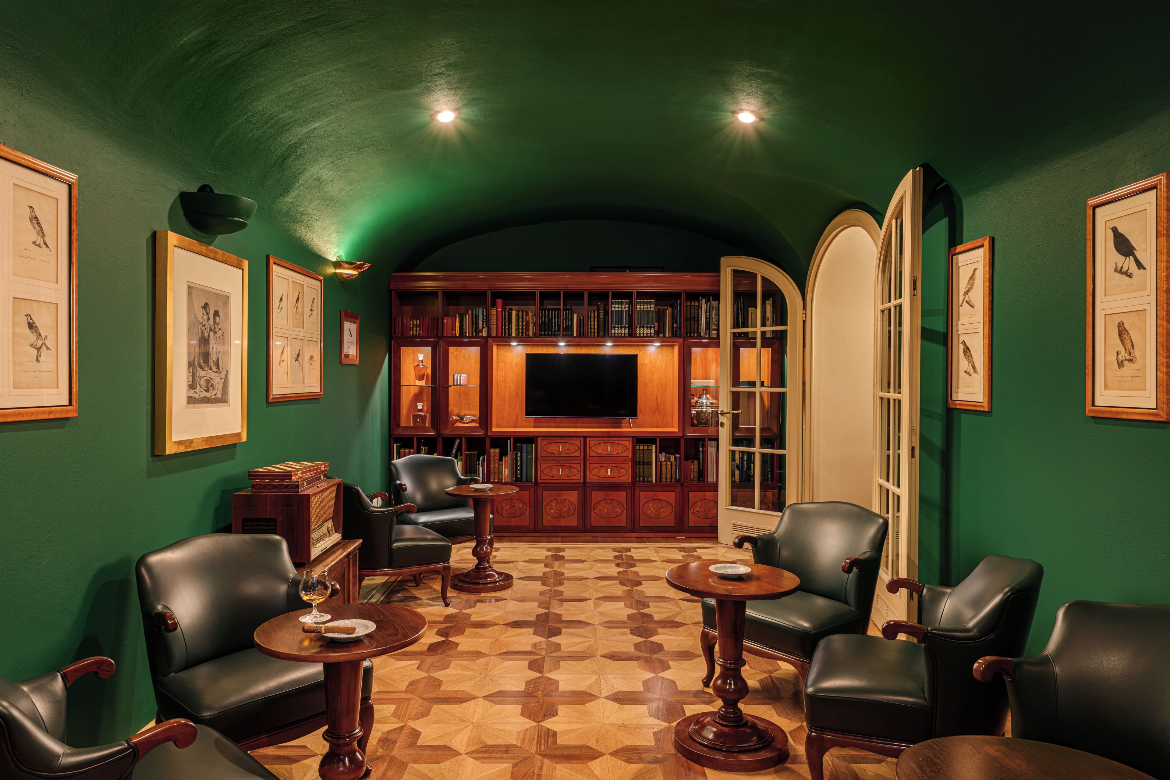 Een kamer met een groene wand, een boekenkast, een televisietoestel, leren fauteuils en een vloer met patroon.