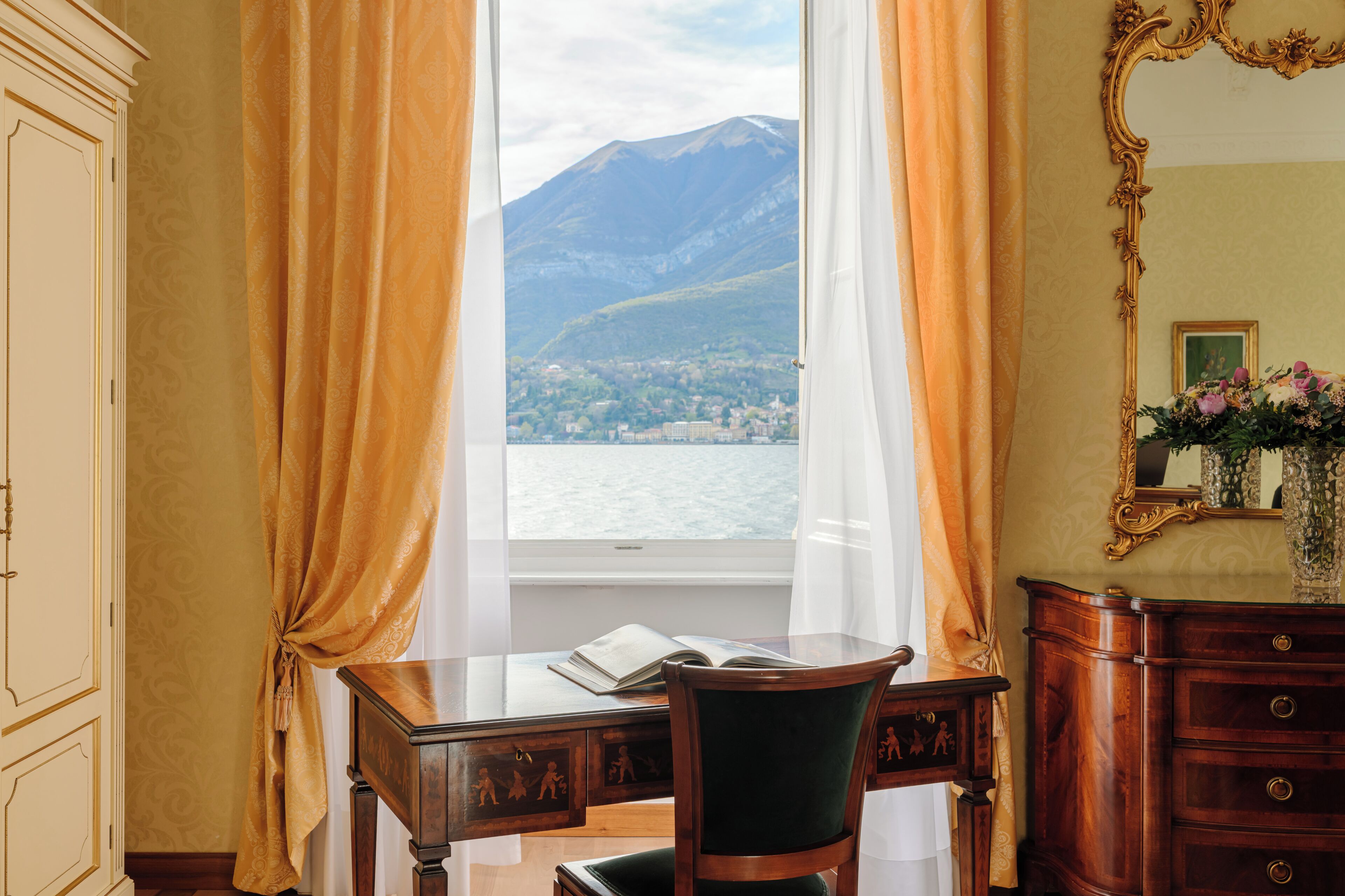A room with a desk, chair, and a view of a lake and mountains through a window.