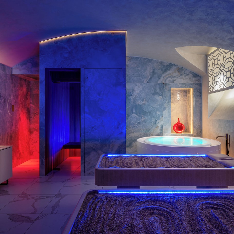 A modern bathroom with a marble wall, a round mirror, a white cabinet, a bathtub with blue LED lights, and a red light fixture.