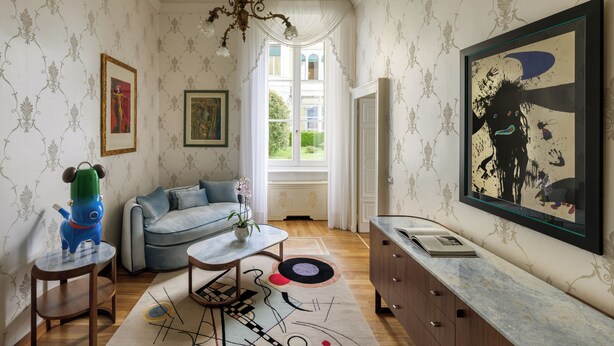 Een kamer met een bank, een bijzettafeltje, een decoratief beeld, een schilderij en een raam met uitzicht op groen.