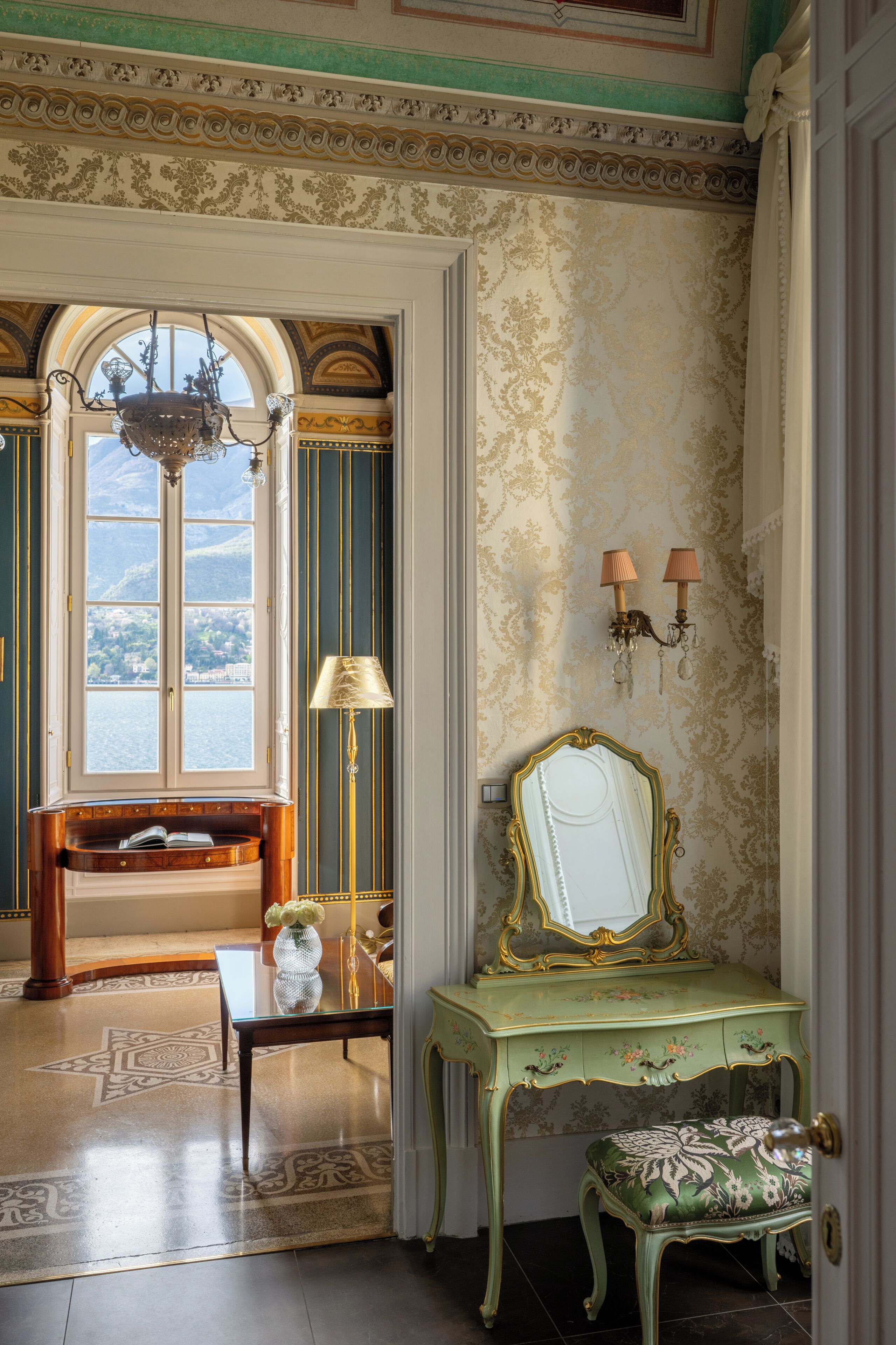 Una habitación con vista al exterior por una ventana, un espejo, una lámpara y un banco.