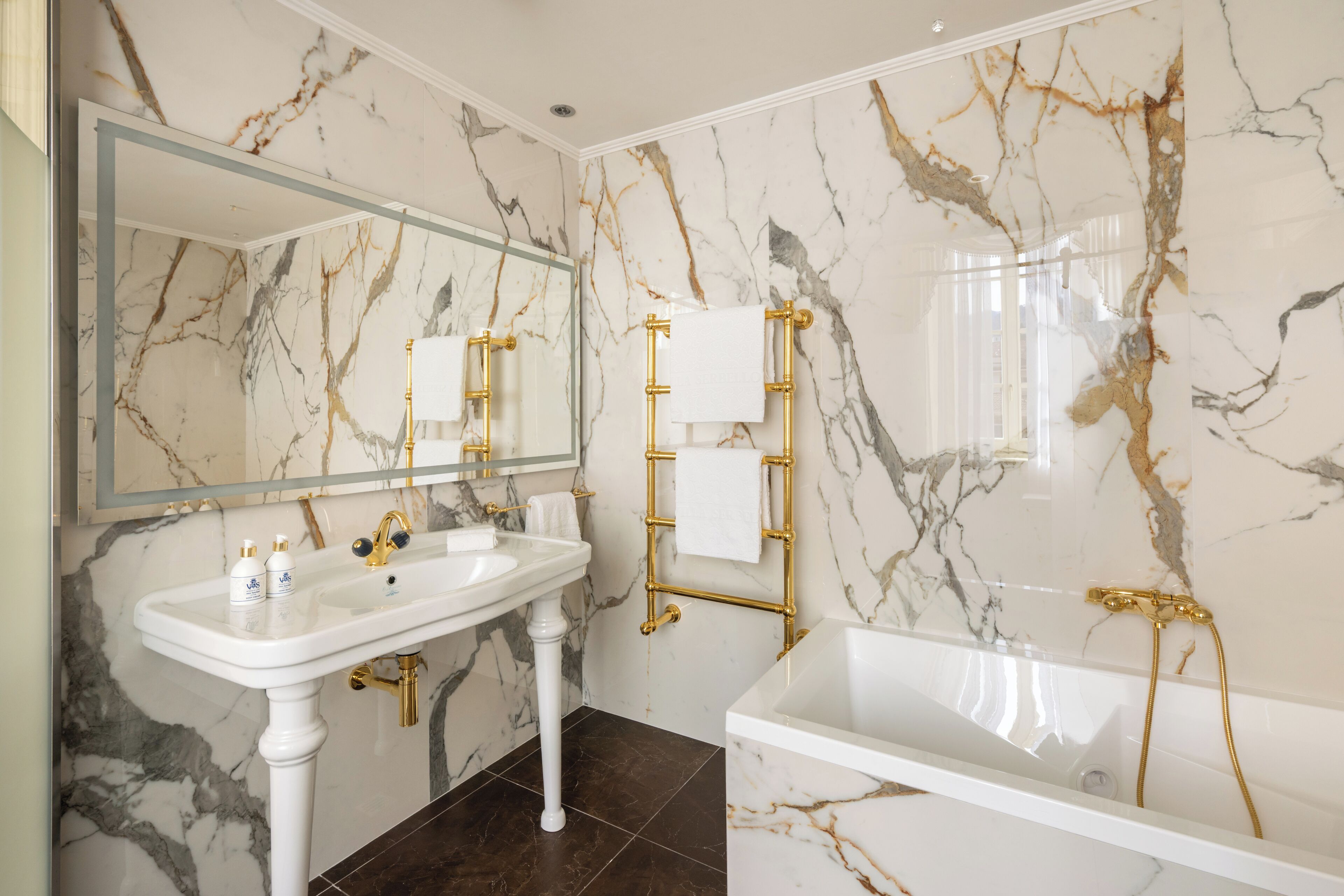 Un baño con pared de mármol, lavabo blanco, espejo y bañera con detalles dorados.