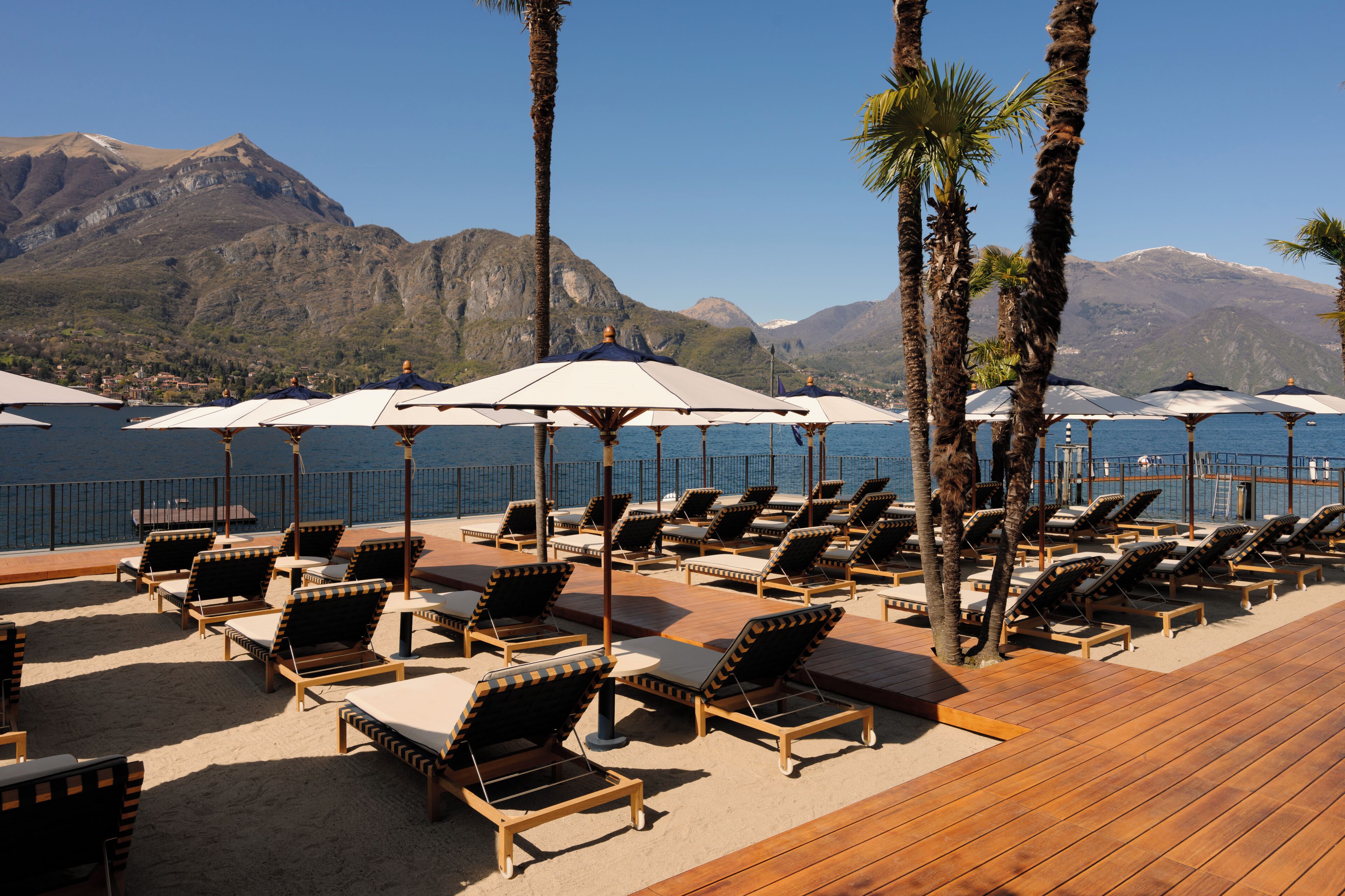 A deck with sun loungers and umbrellas, overlooking a body of water and mountains.