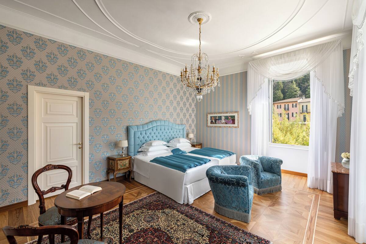 Une chambre à coucher avec un grand lit, un lustre, une vue sur un immeuble par la fenêtre et un tapis à motifs.