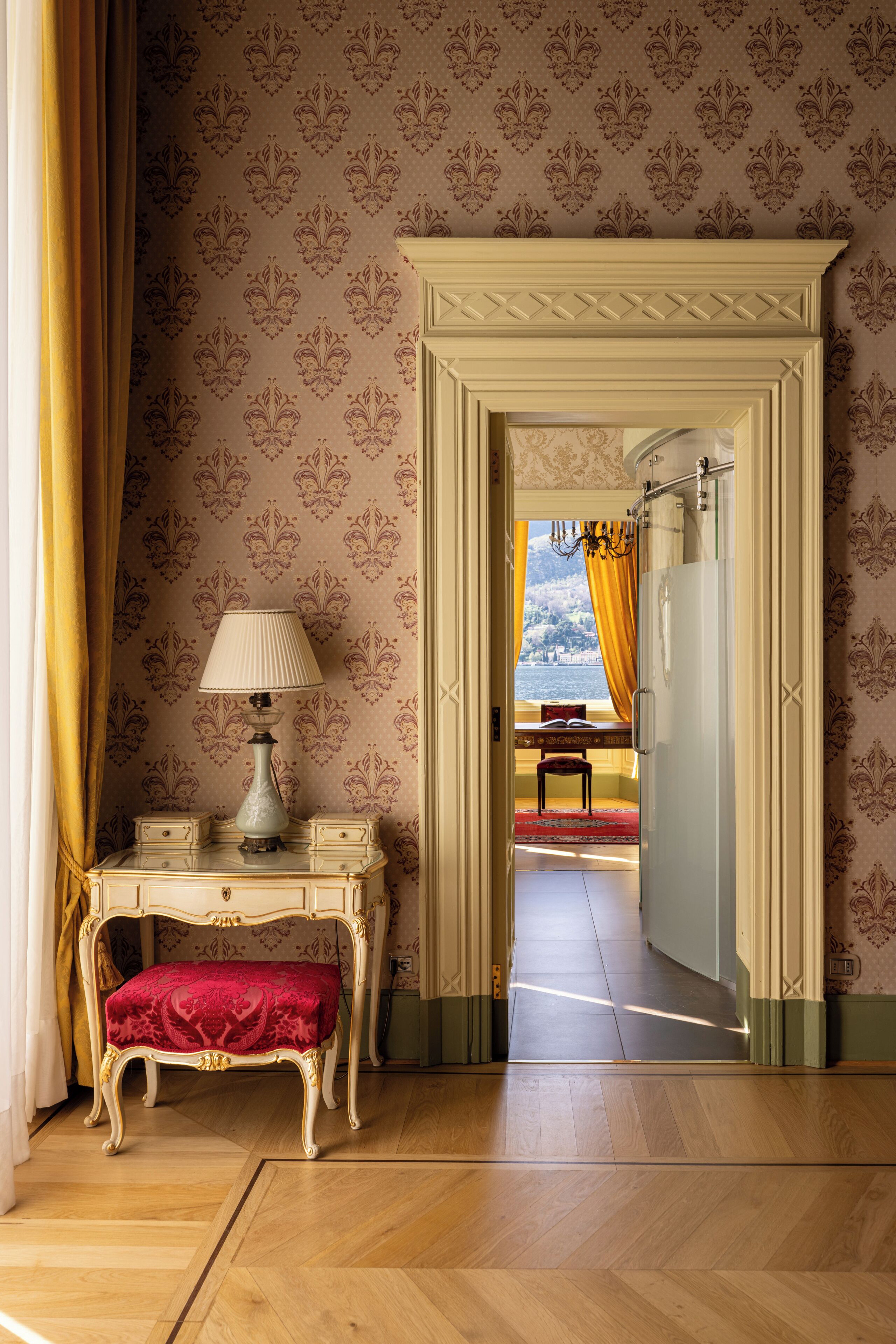 A room with a wooden floor, a red upholstered bench, a white table with a lamp, and a doorway leading to another room with a view.