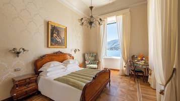 Ein Schlafzimmer mit einem hölzernen Bett, einem Gemälde an der Wand, einer Chandelierleuchtre, einem Sessel und einem Schreibtisch mit Blick ins Freie.