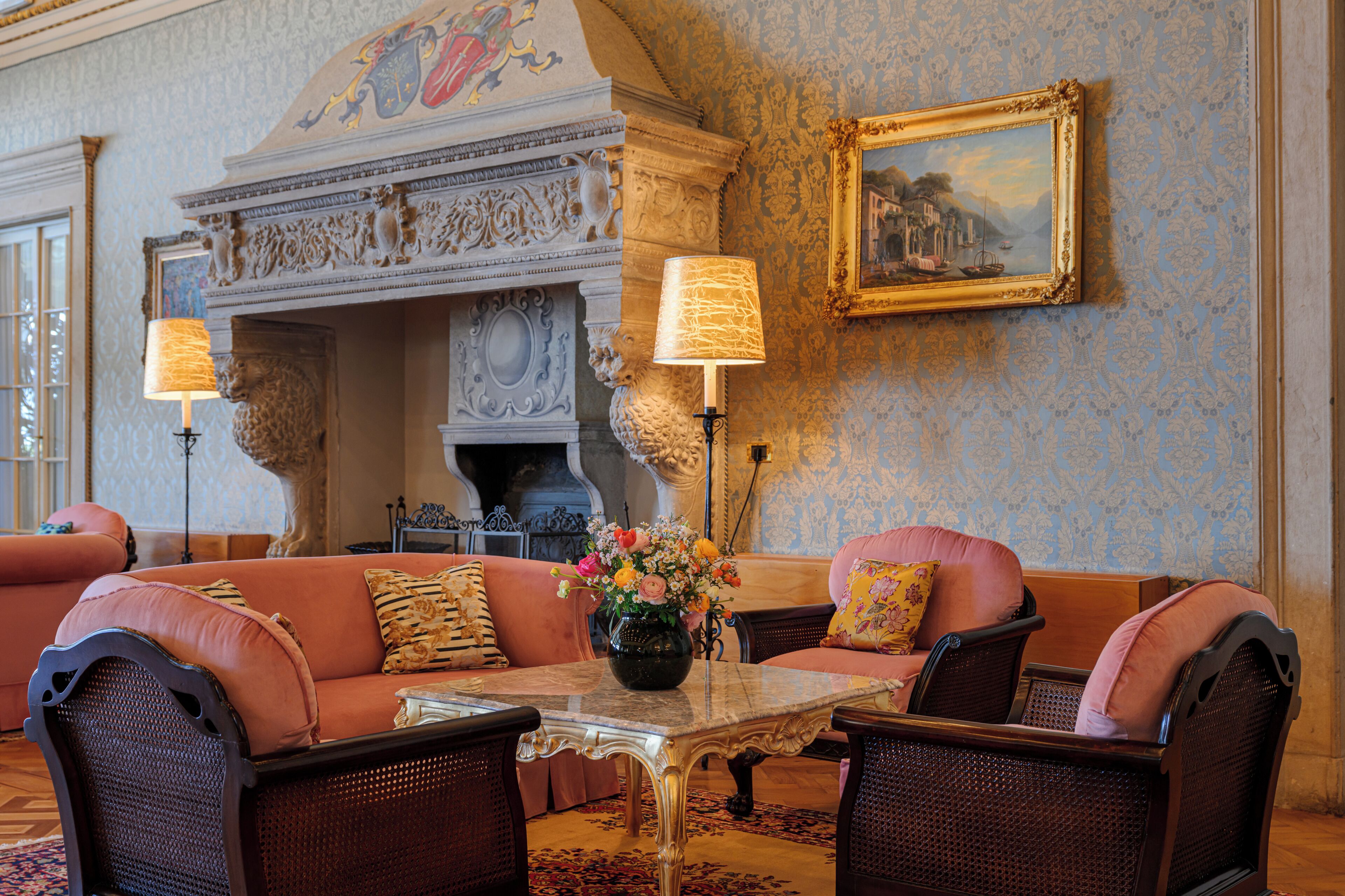 A room with a fireplace, a sofa set, a table with a vase of flowers, and a framed painting on the wall.