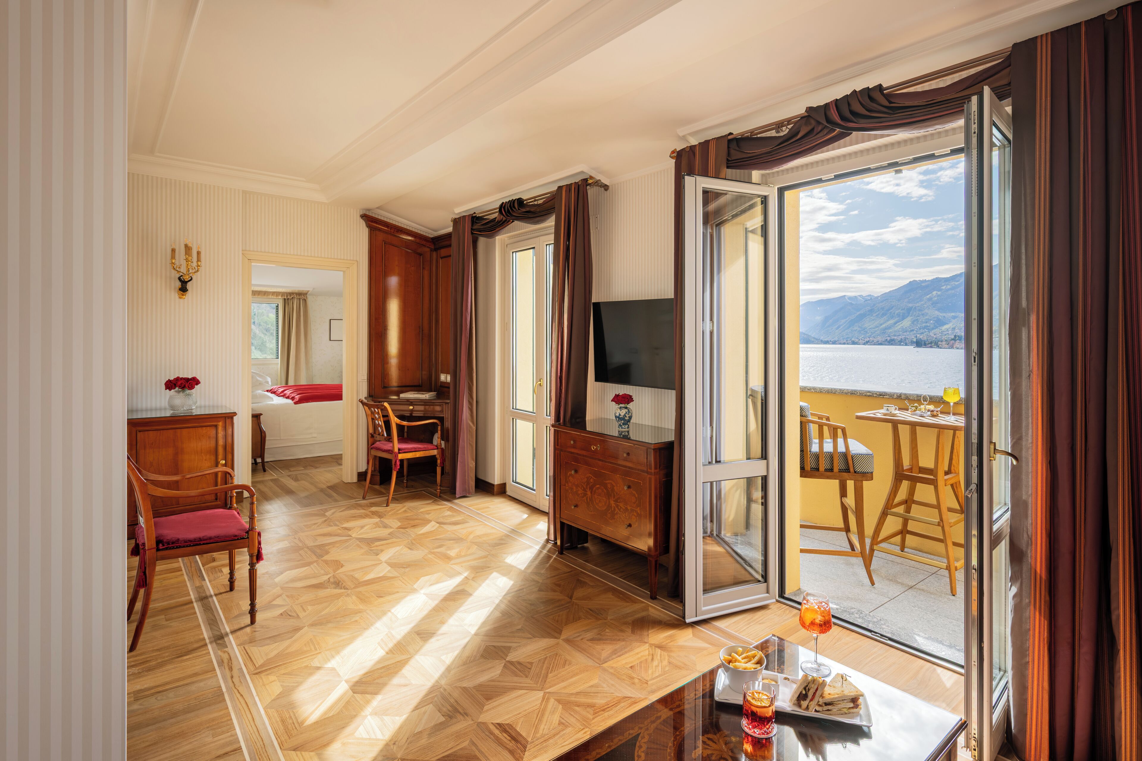 Une chambre d’hôtel avec un parquet en bois, un coin repas comprenant une table et des chaises, une télévision, et une vue sur un lac et des montagnes, visible par la porte ouverte.