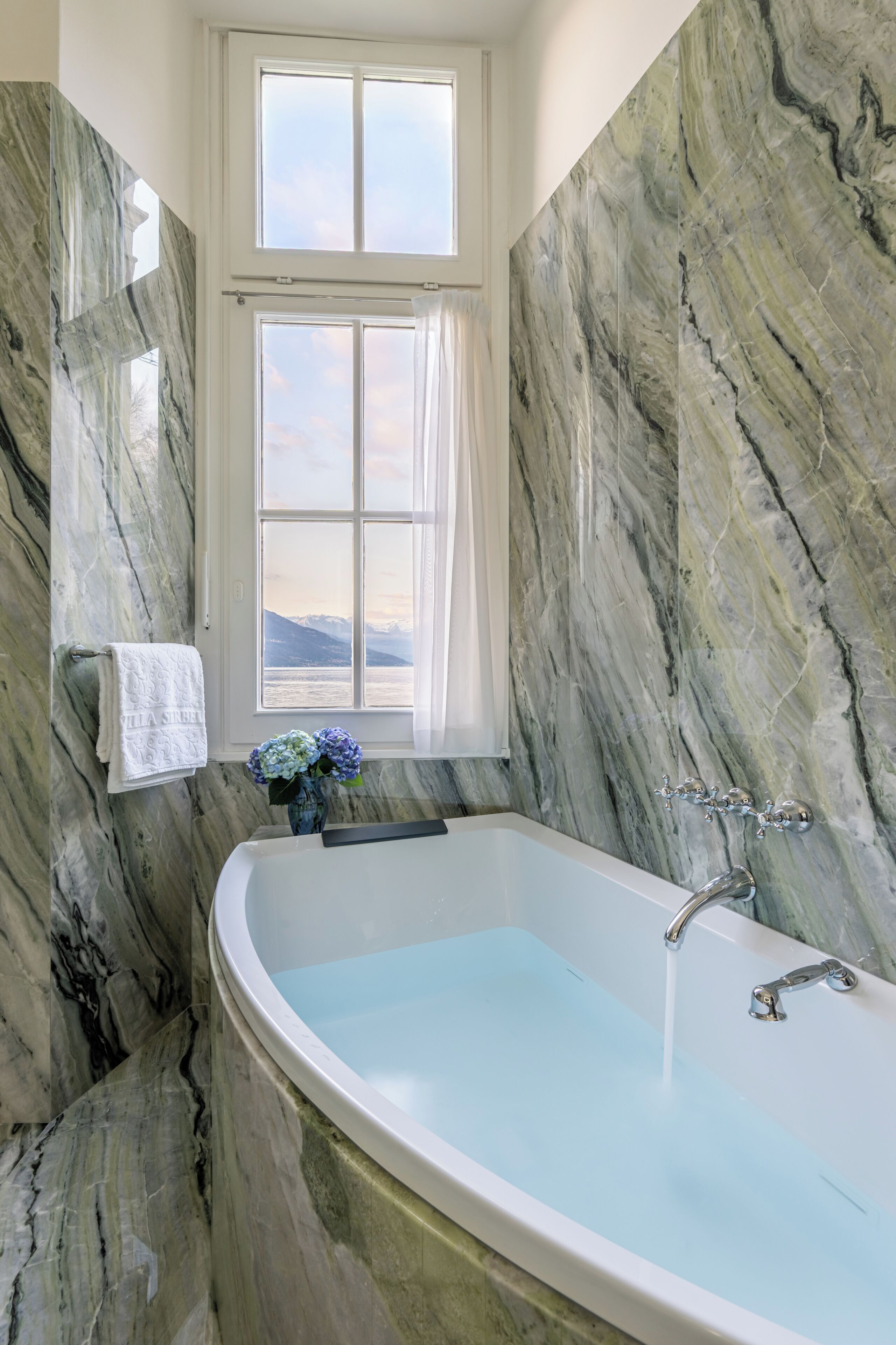 Ein Badezimmer mit einer großen Badewanne, Marmorwänden, einem Fenster mit Blick und einem Handtuchhalter.