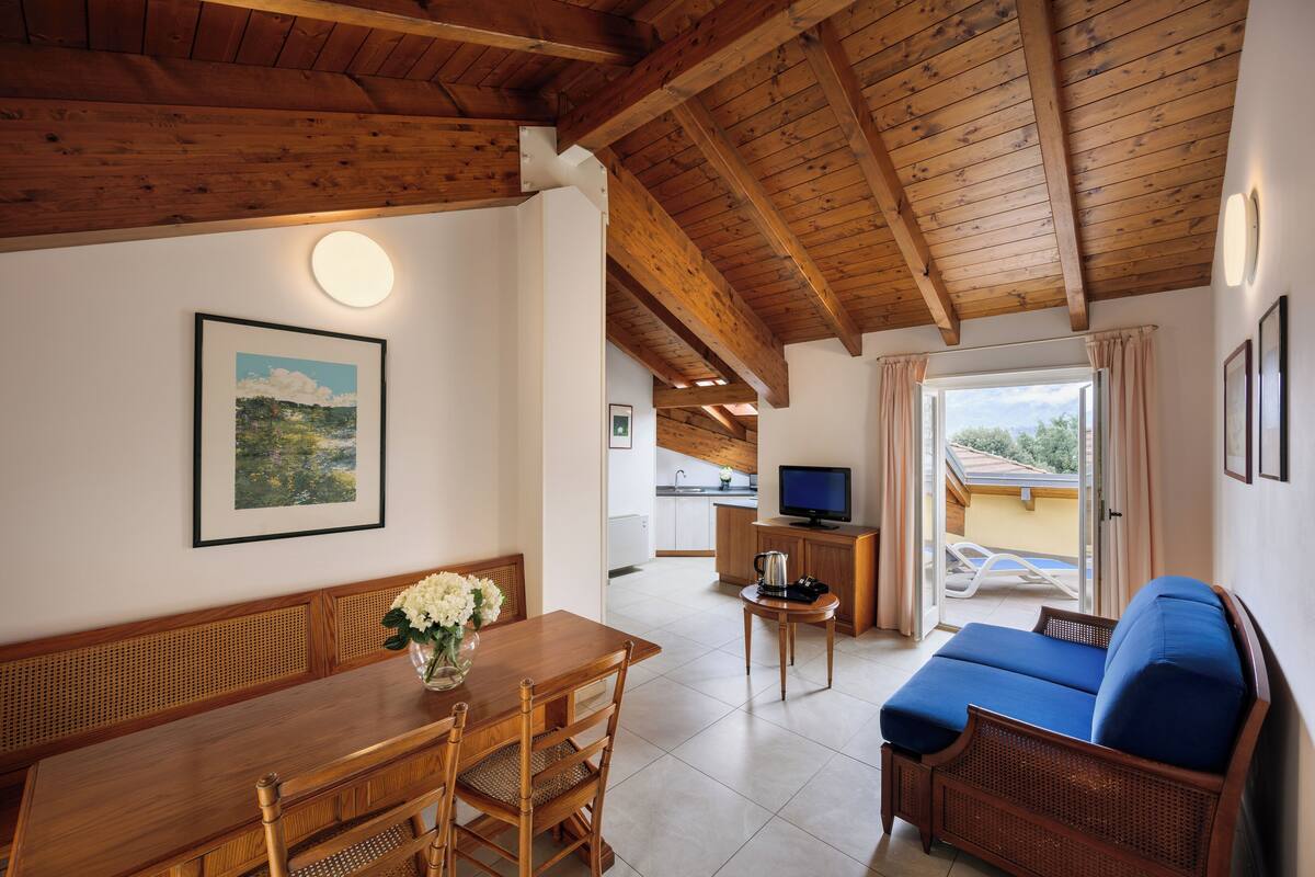 A cozy living area with a wooden ceiling, a dining table with chairs, a sofa, and a television on a stand.