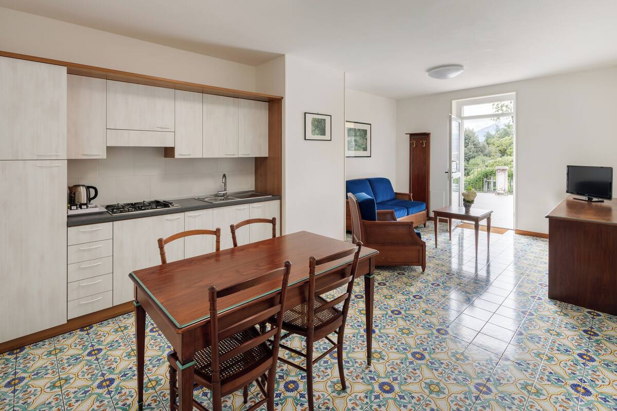 A modern kitchen with white cabinets and a dining area with a wooden table and chairs. A blue sofa and a TV are visible in the living area.