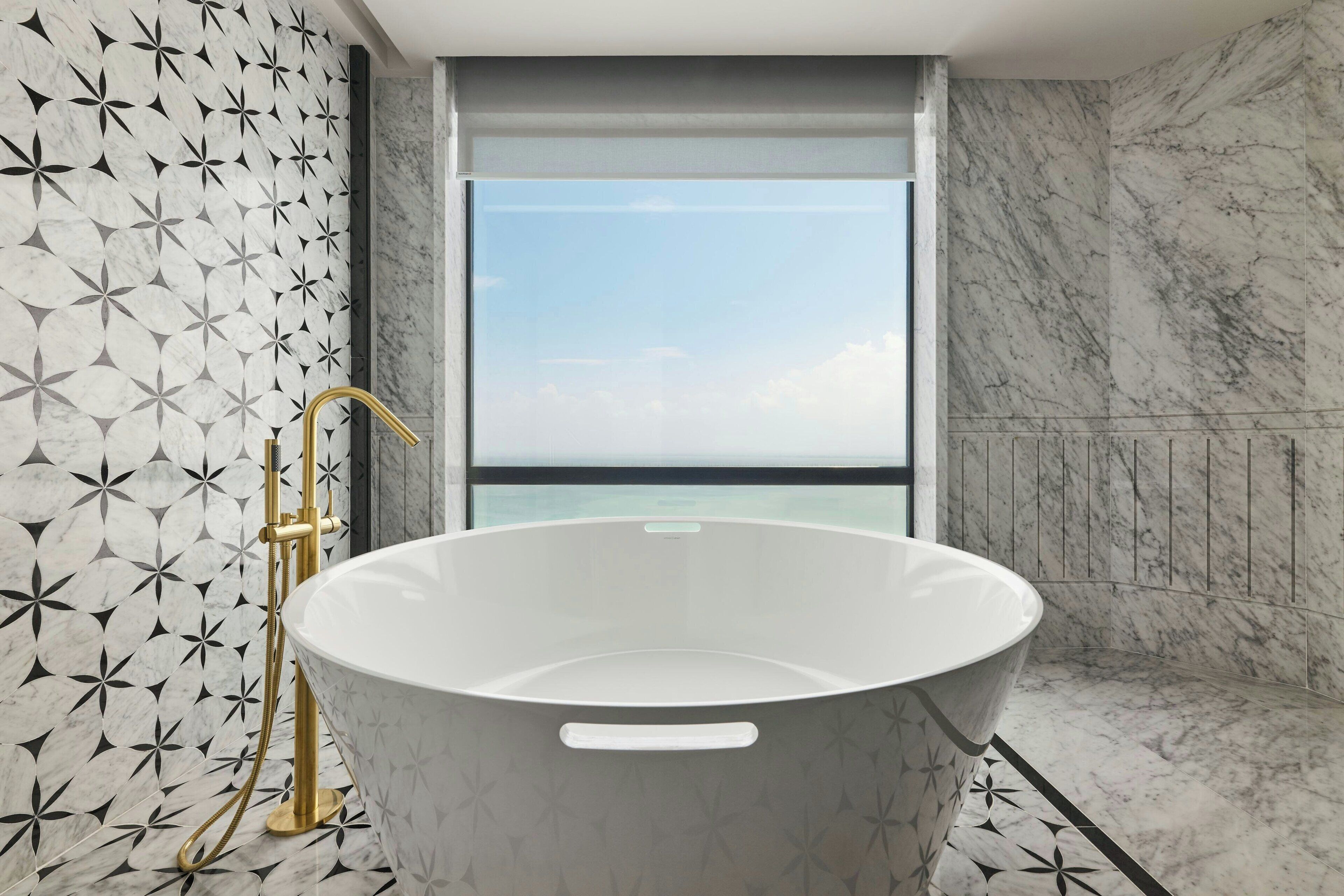 Ein modernes Badezimmer mit einer freistehenden Badewanne, einer Dusche mit goldfarbener Armatur und einem großen Fenster mit Blick auf das Meer.