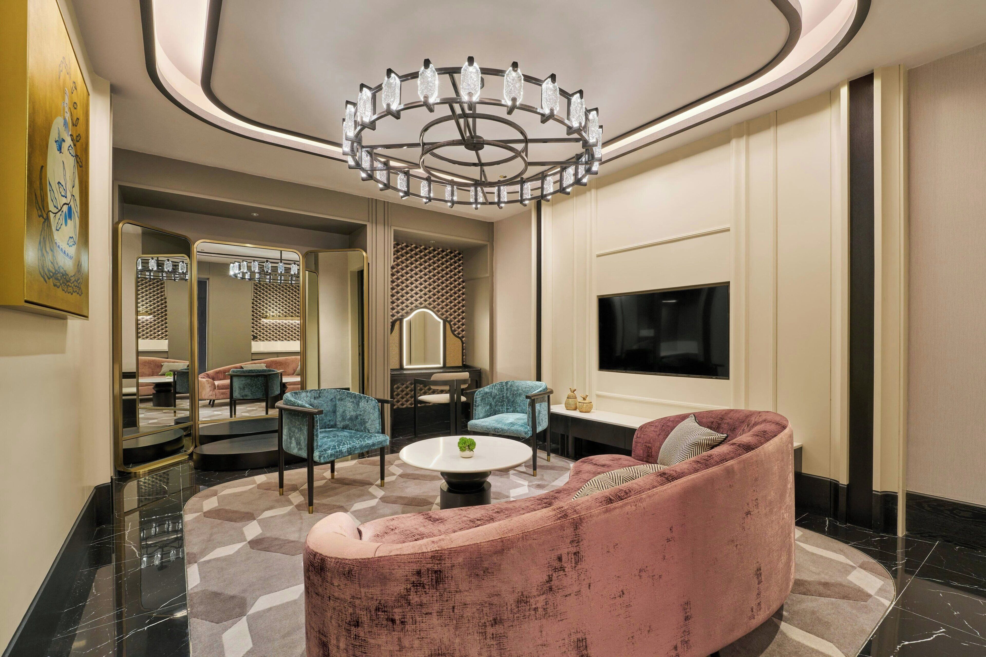 A modern living room with a pink sofa, blue chairs, a round coffee table, and a large TV on the wall.