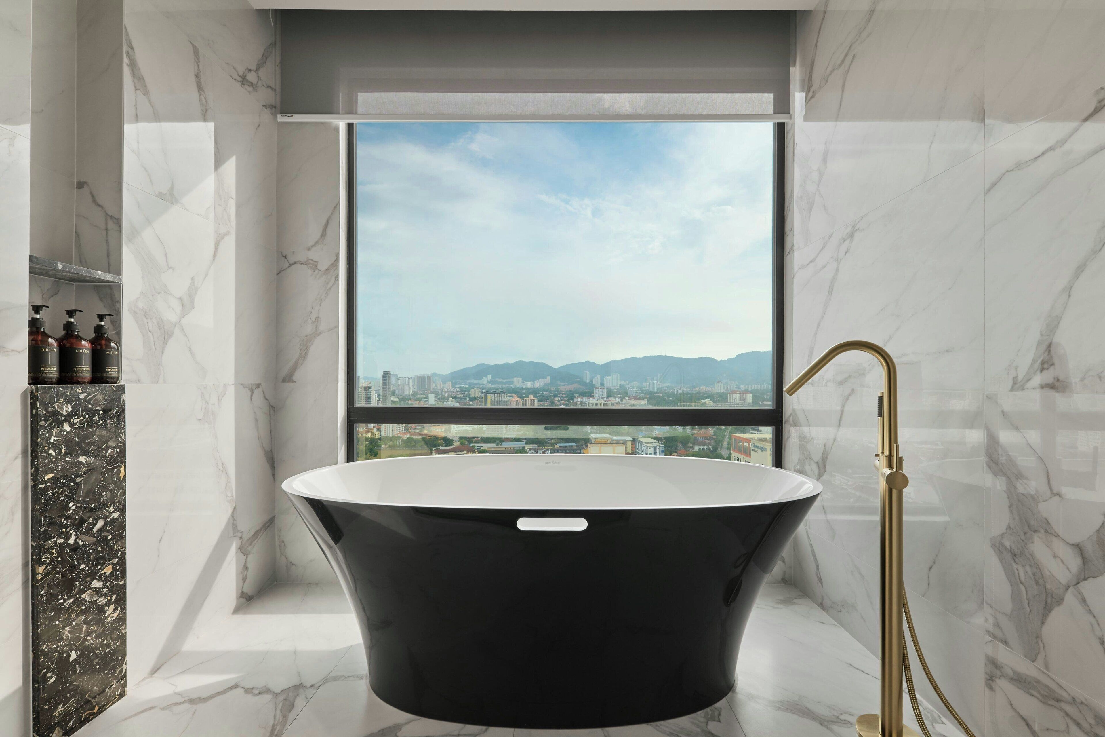 A modern bathroom with a freestanding bathtub, marble walls, and a city view through a large window.