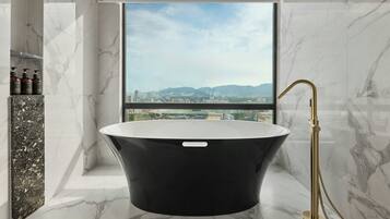 A modern bathroom with a freestanding bathtub, marble walls, and a city view through a large window.