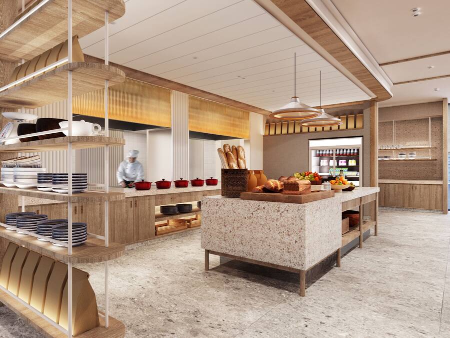 Une cuisine moderne avec des meubles en bois, un îlot central agrémenté de fruits et de pain, et un chef qui prépare les plats.