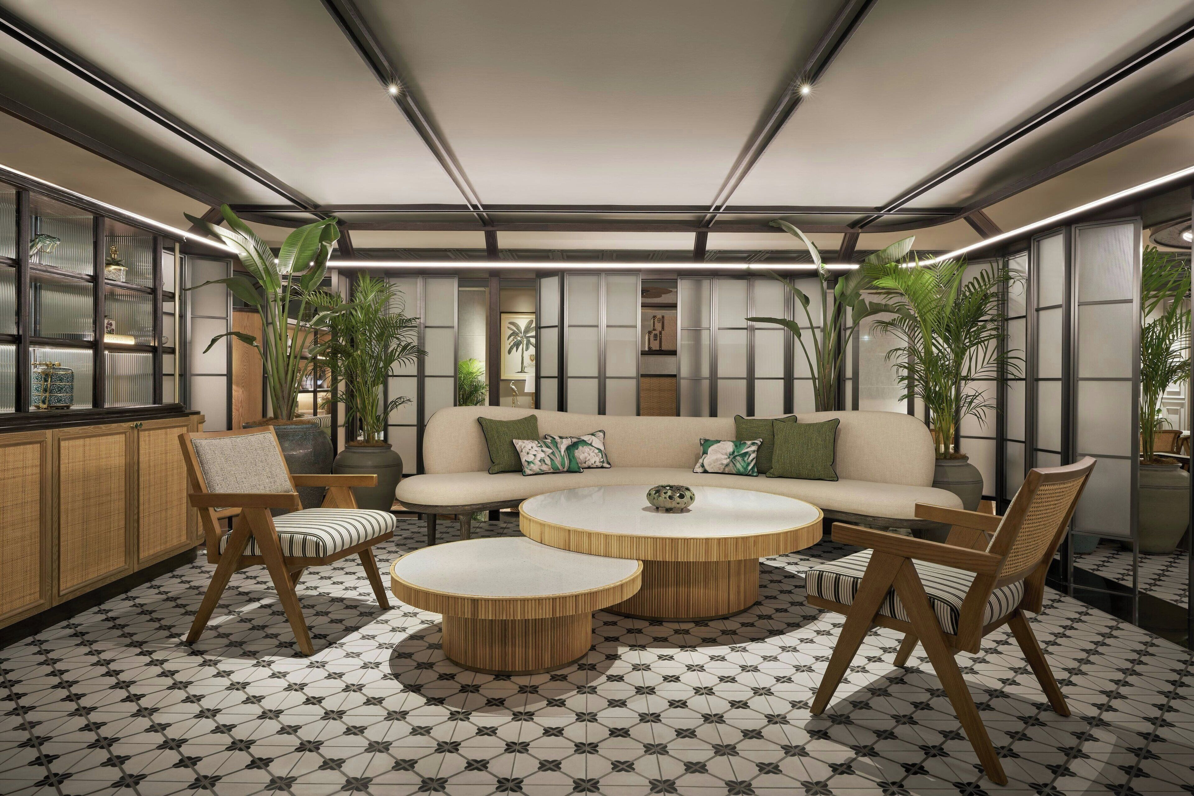 Een moderne woonruimte met een bank, stoelen en ronde tafels, met een vloer met geometrisch patroon en grote ramen met planten.