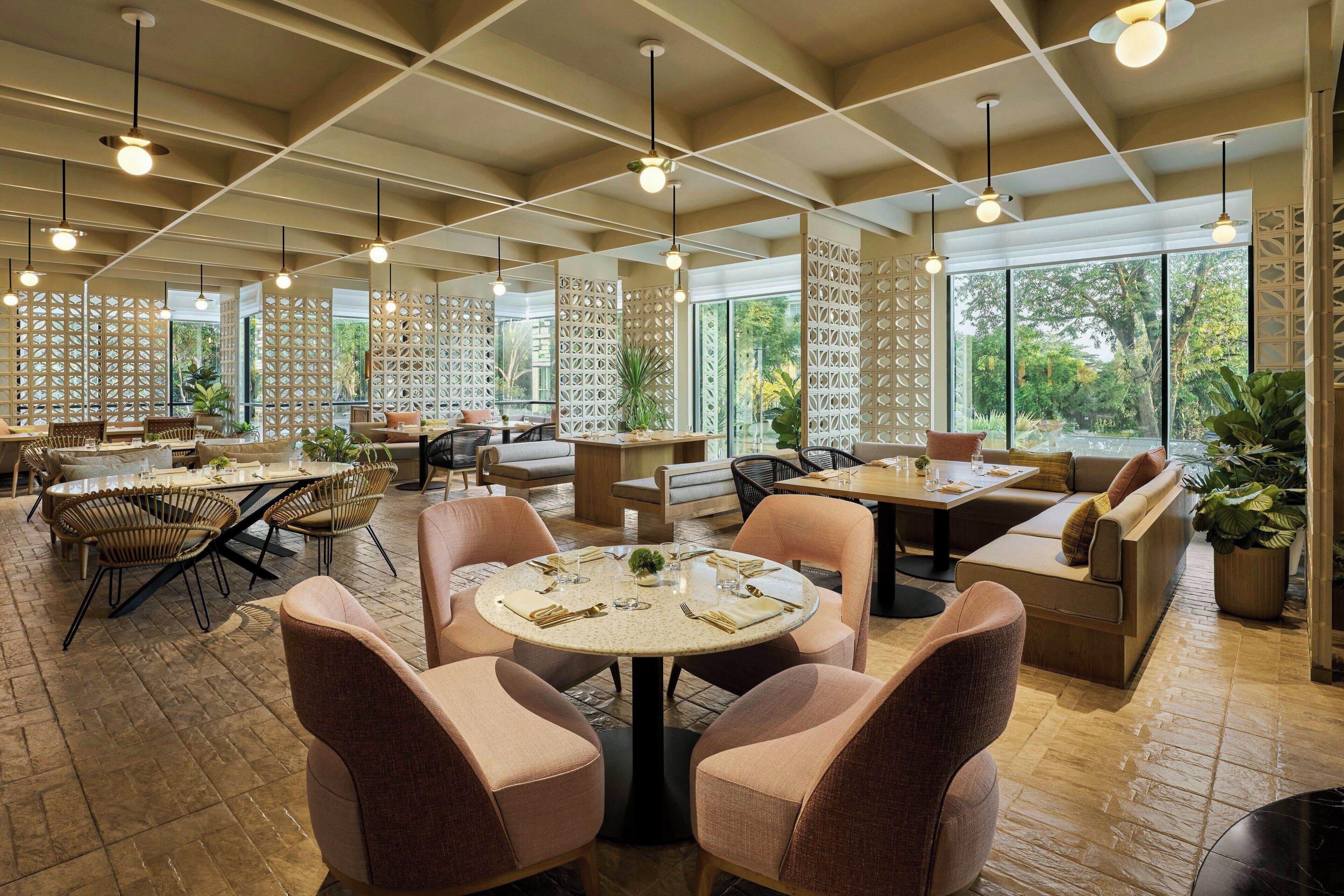 A spacious restaurant with round tables and cushioned seating, large windows offering a view of greenery, and pendant lighting.