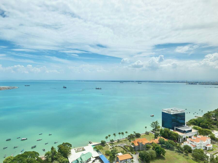 An aerial view of a coastal area with clear blue waters, boats, and buildings including a prominent modern structure.