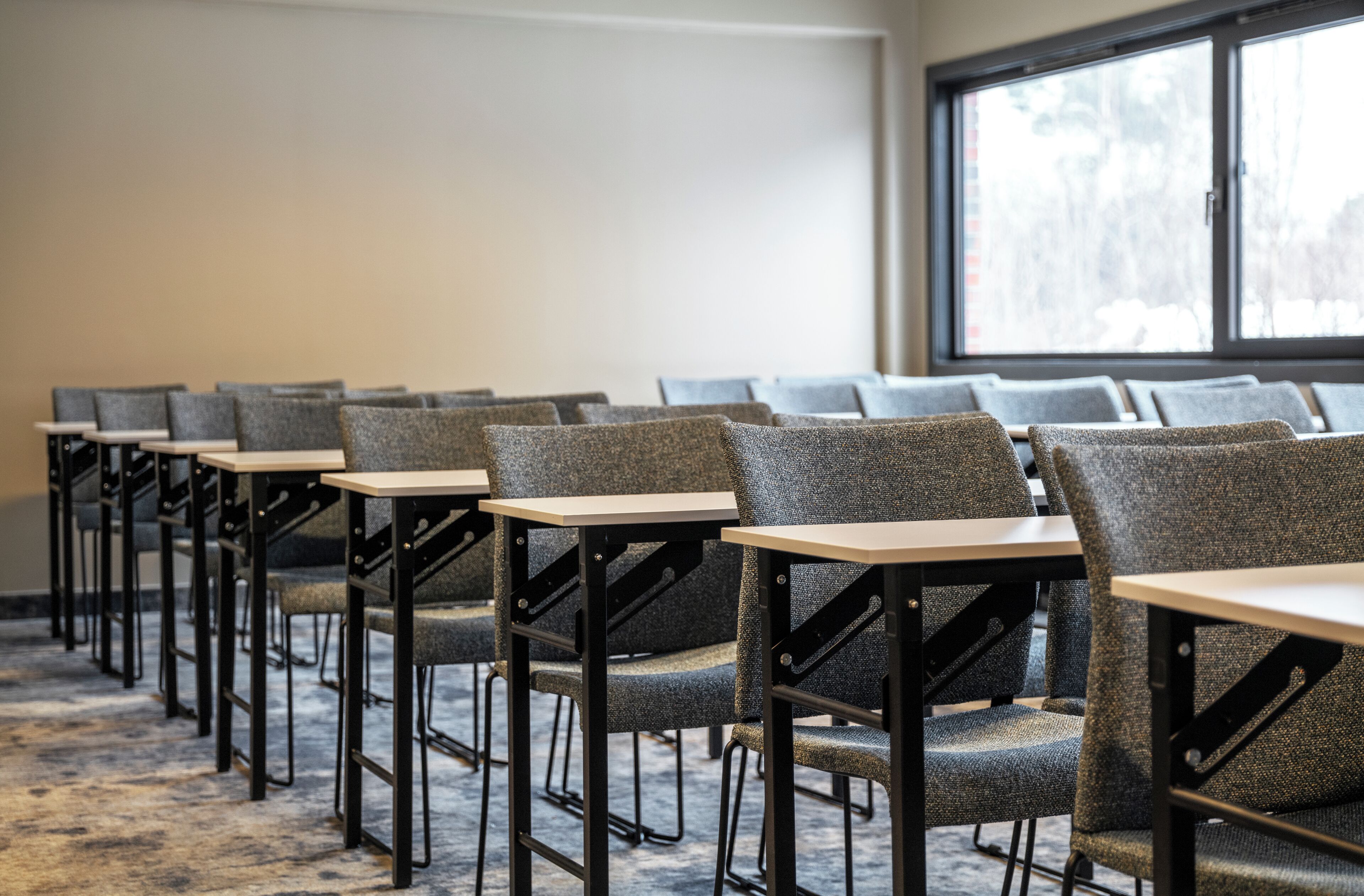 A row of grey upholstered chairs with black metal frames and wooden tables, arranged in a conference room with a large window in the background.