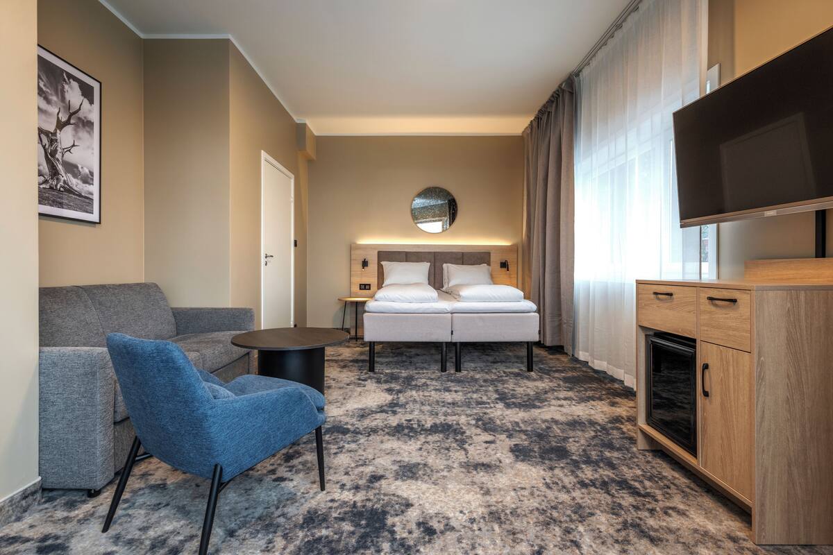 A hotel room with a bed, a blue armchair, a small table, a TV, and a wooden cabinet.