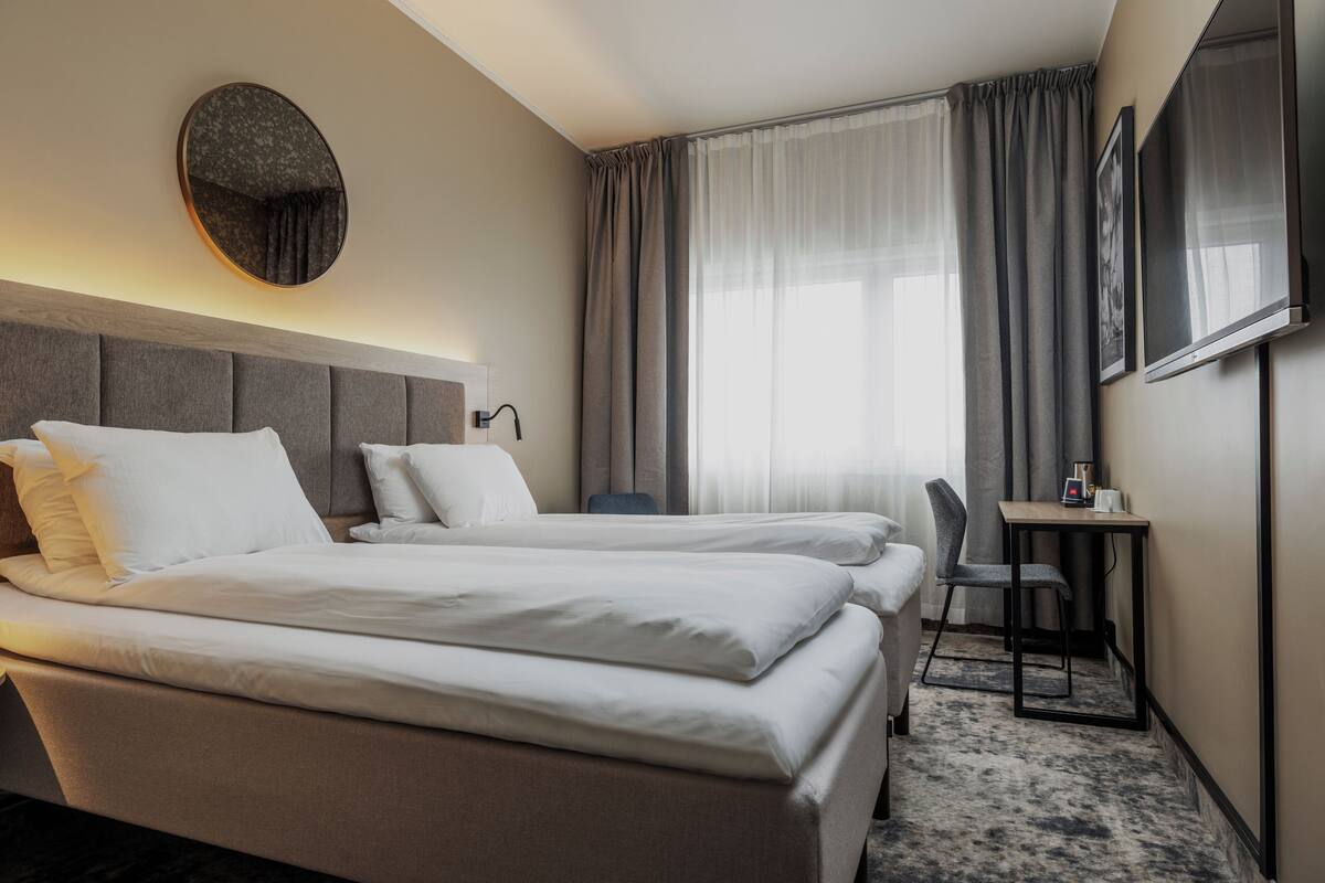 A hotel room with two beds, a desk, a chair, a TV, and a large window with curtains.