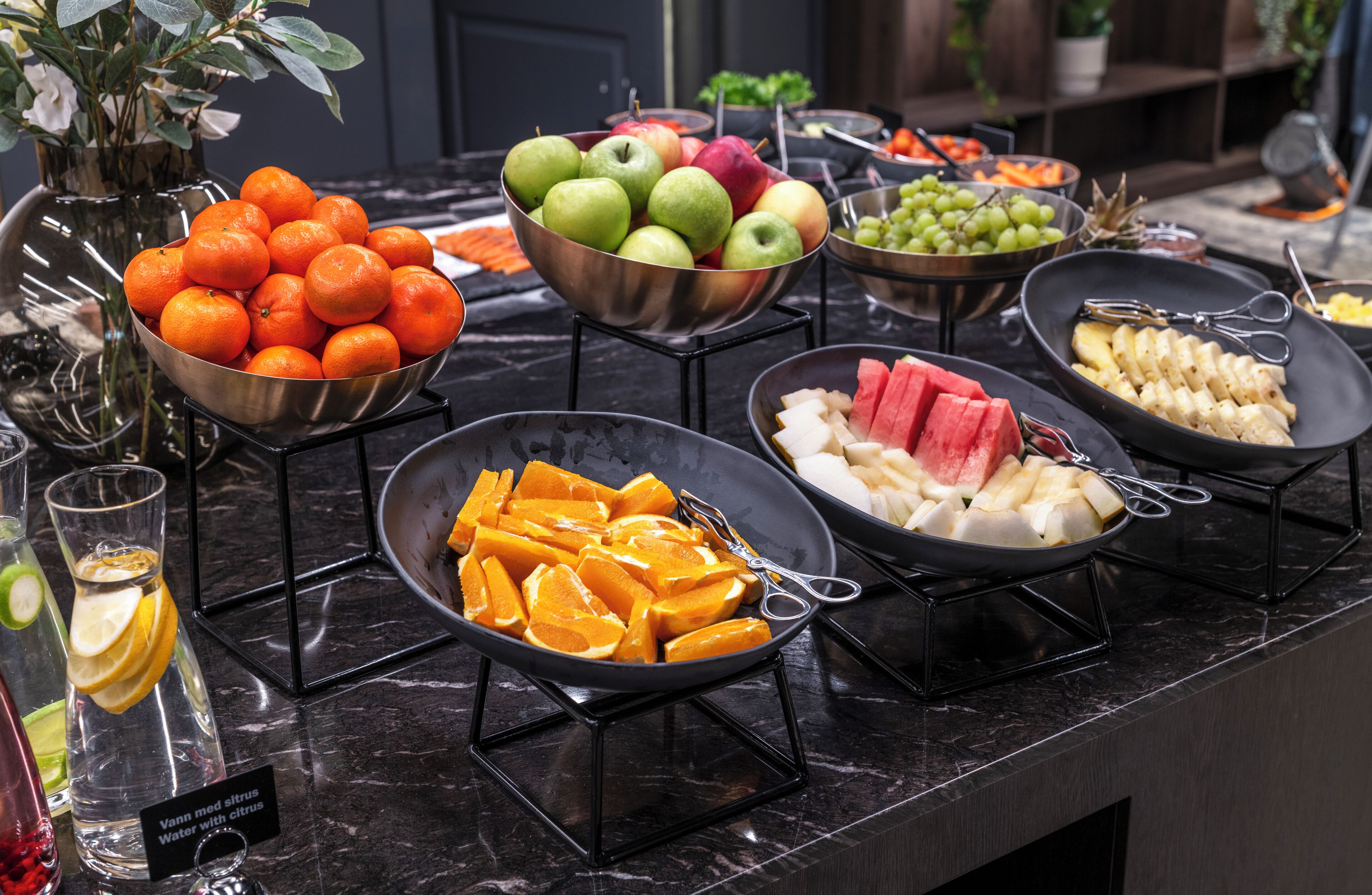 A buffet table with various fruits and vegetables, including oranges, apples, and watermelon, arranged on a marble countertop.