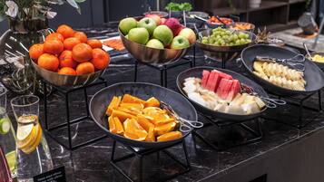 A buffet table with various fruits and vegetables, including oranges, apples, and watermelon, arranged on a marble countertop.