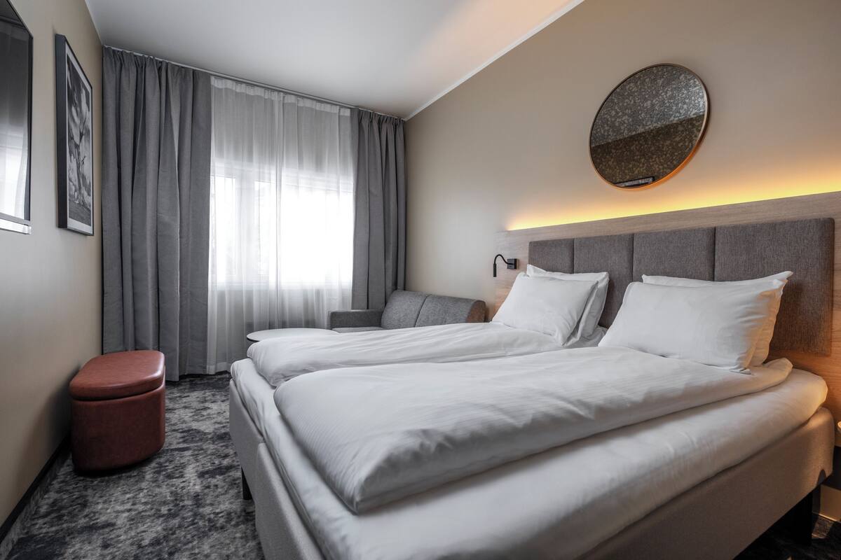 A hotel room with two beds, a round mirror, a nightstand, and a red ottoman.
