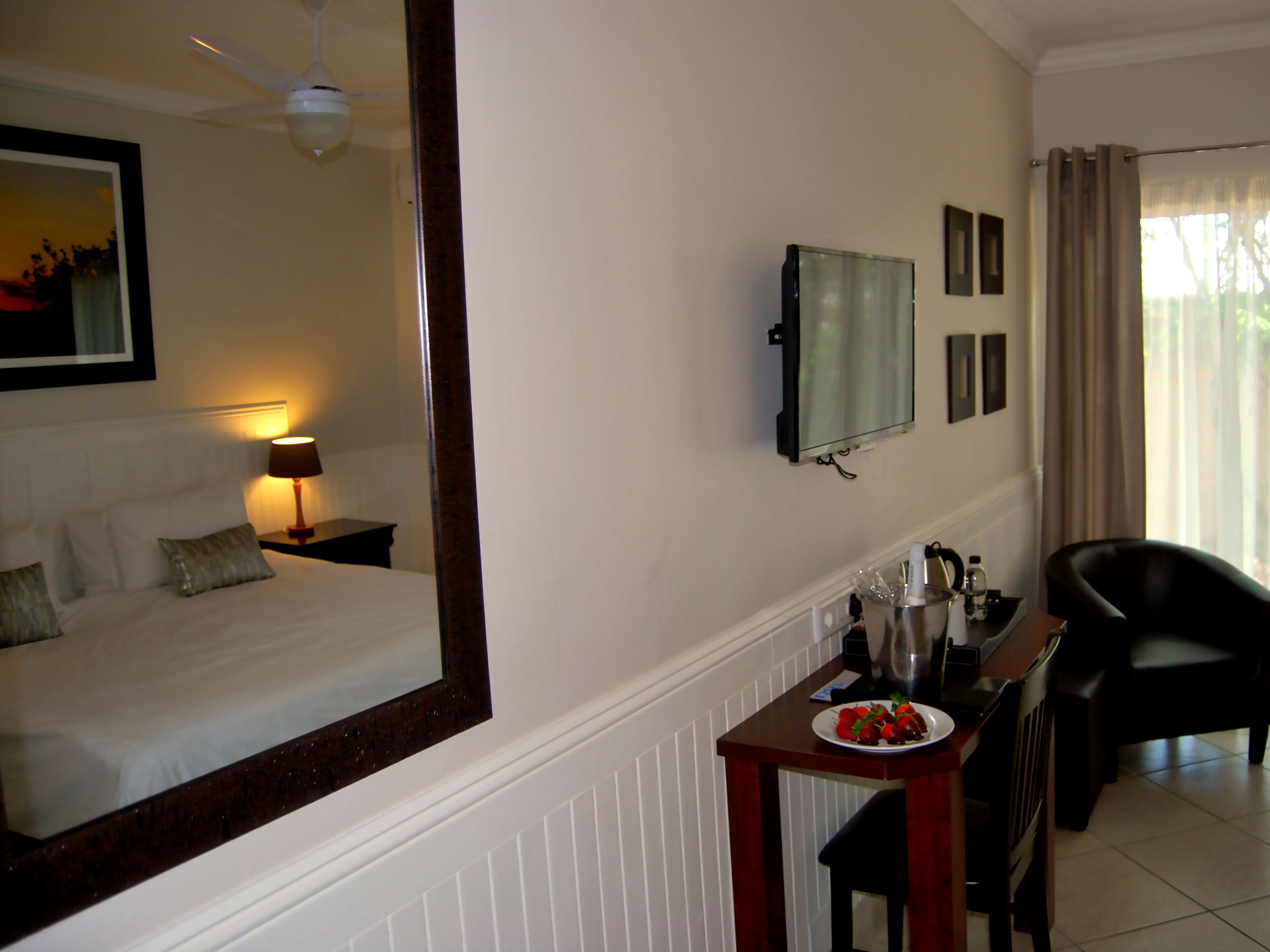 A hotel room with a bed, a desk with a plate of strawberries, a chair, a TV, and a ceiling fan.