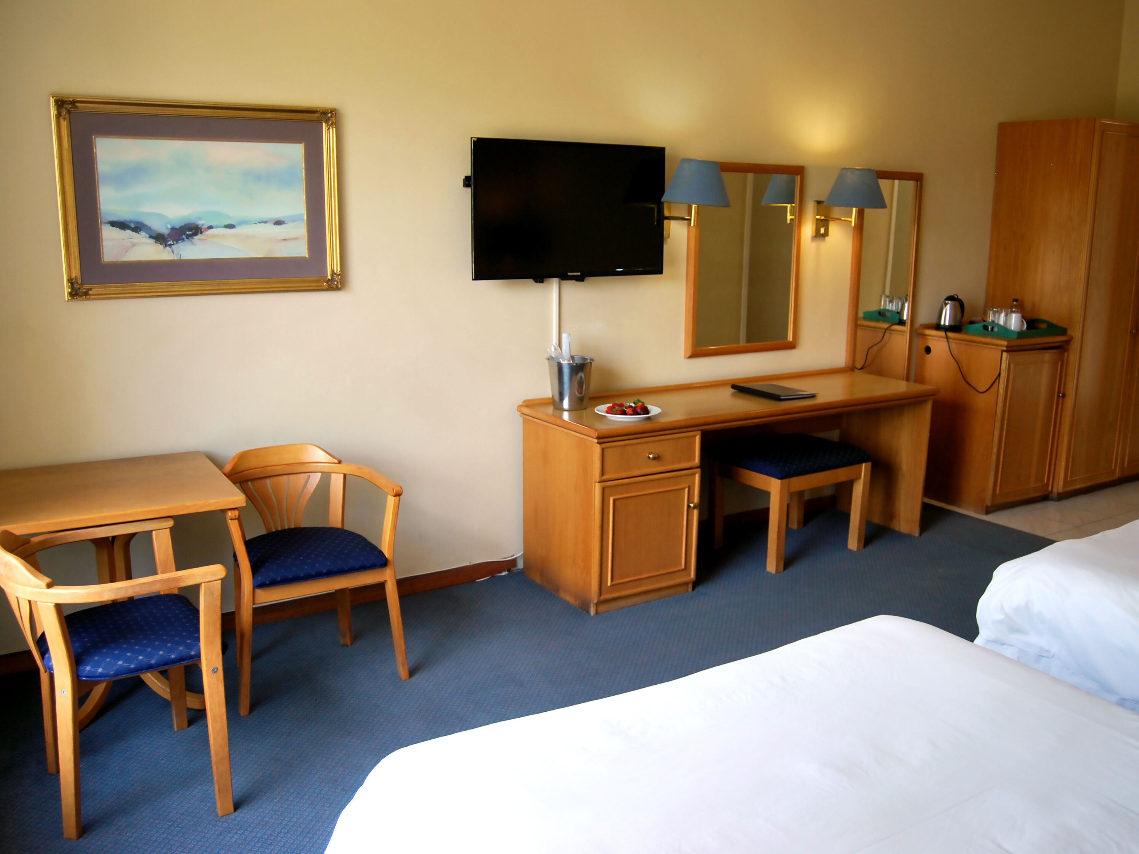 A hotel room with a desk, chair, bed, television, and a painting on the wall.