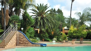 A pool area with a blue slide, palm trees, and a building in the background. - Safari Hotel & Convention Centre (Rustenburg)
