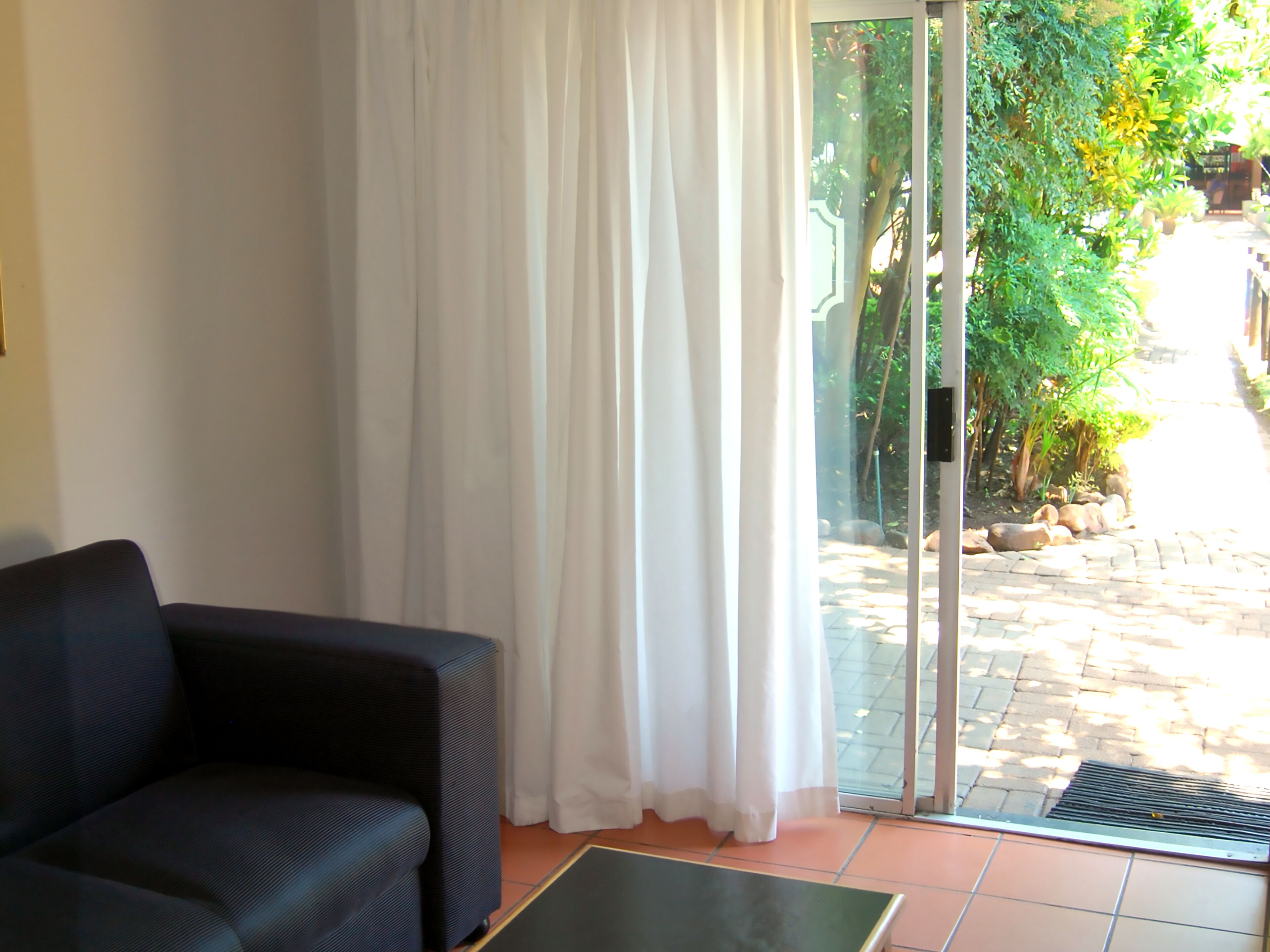 A room with a black sofa, a glass door leading to a garden, and a tiled floor.