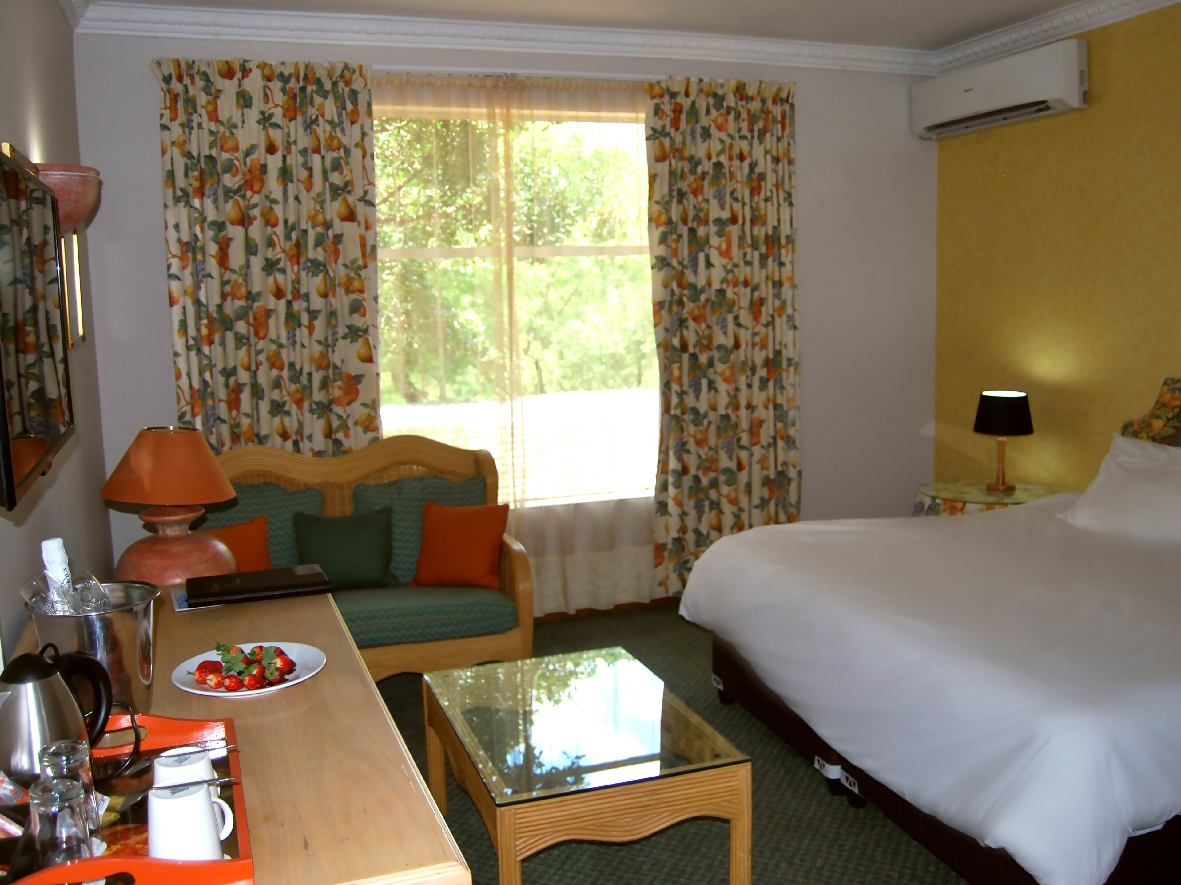 A hotel room with a bed, a sofa, a glass-top table, a desk with a lamp, and a window with curtains.