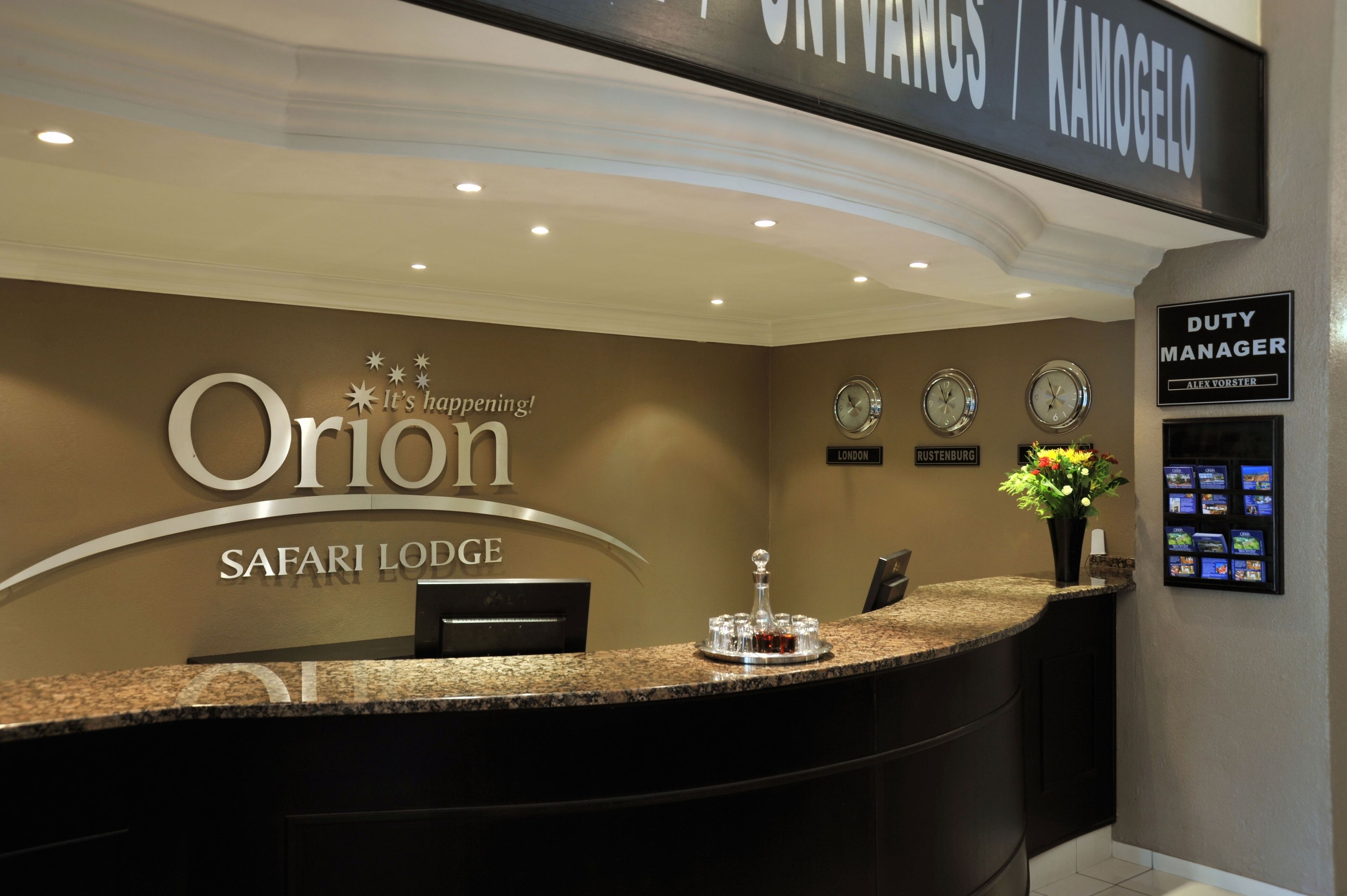 A reception desk at Orion Safari Lodge with a sign above indicating it's happening, clocks showing London and Rustenburg times, and a Duty Manager sign.