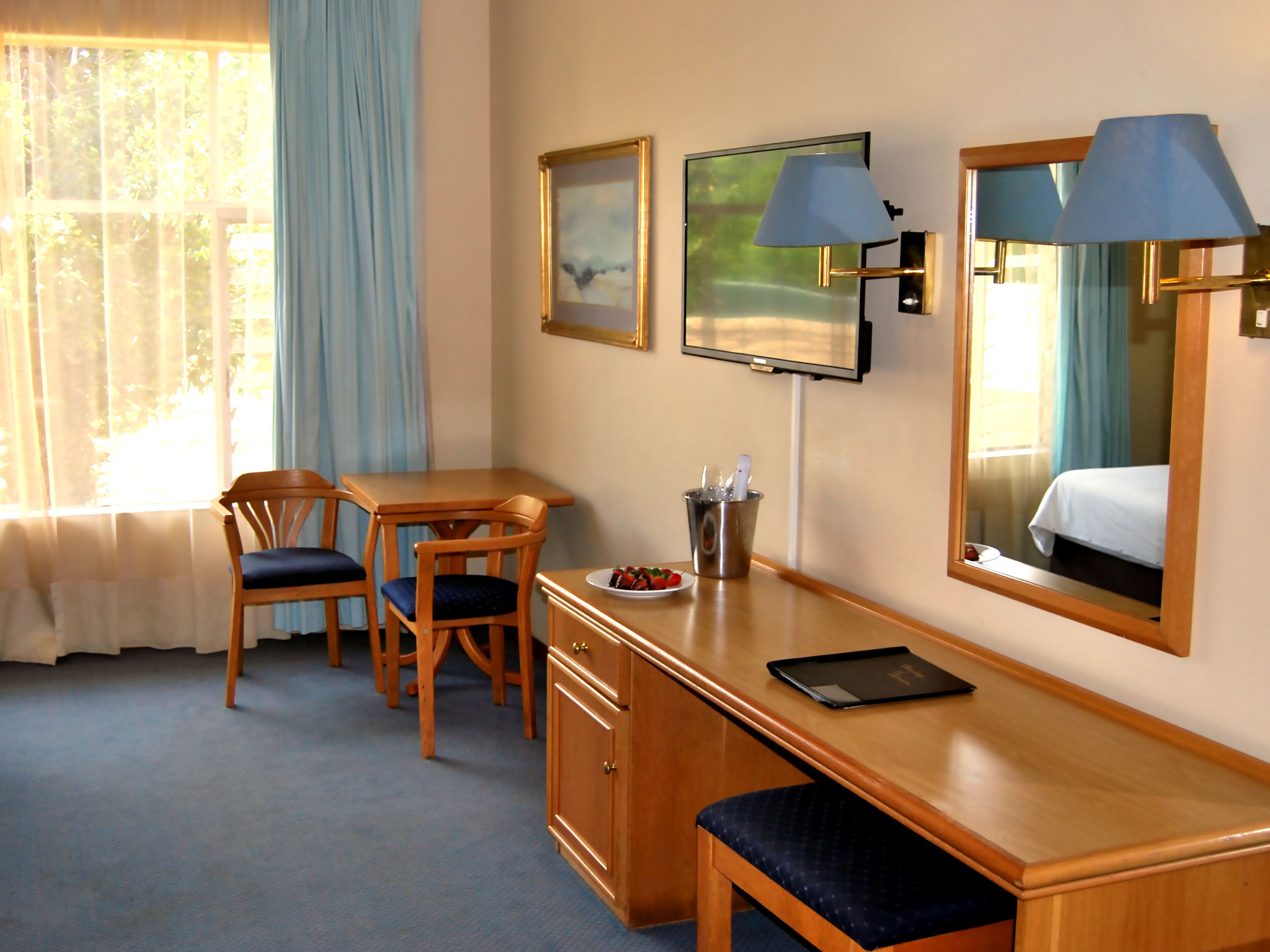 Une chambre d’hôtel avec un bureau en bois, deux chaises, une télévision, un miroir et une fenêtre avec des rideaux.
