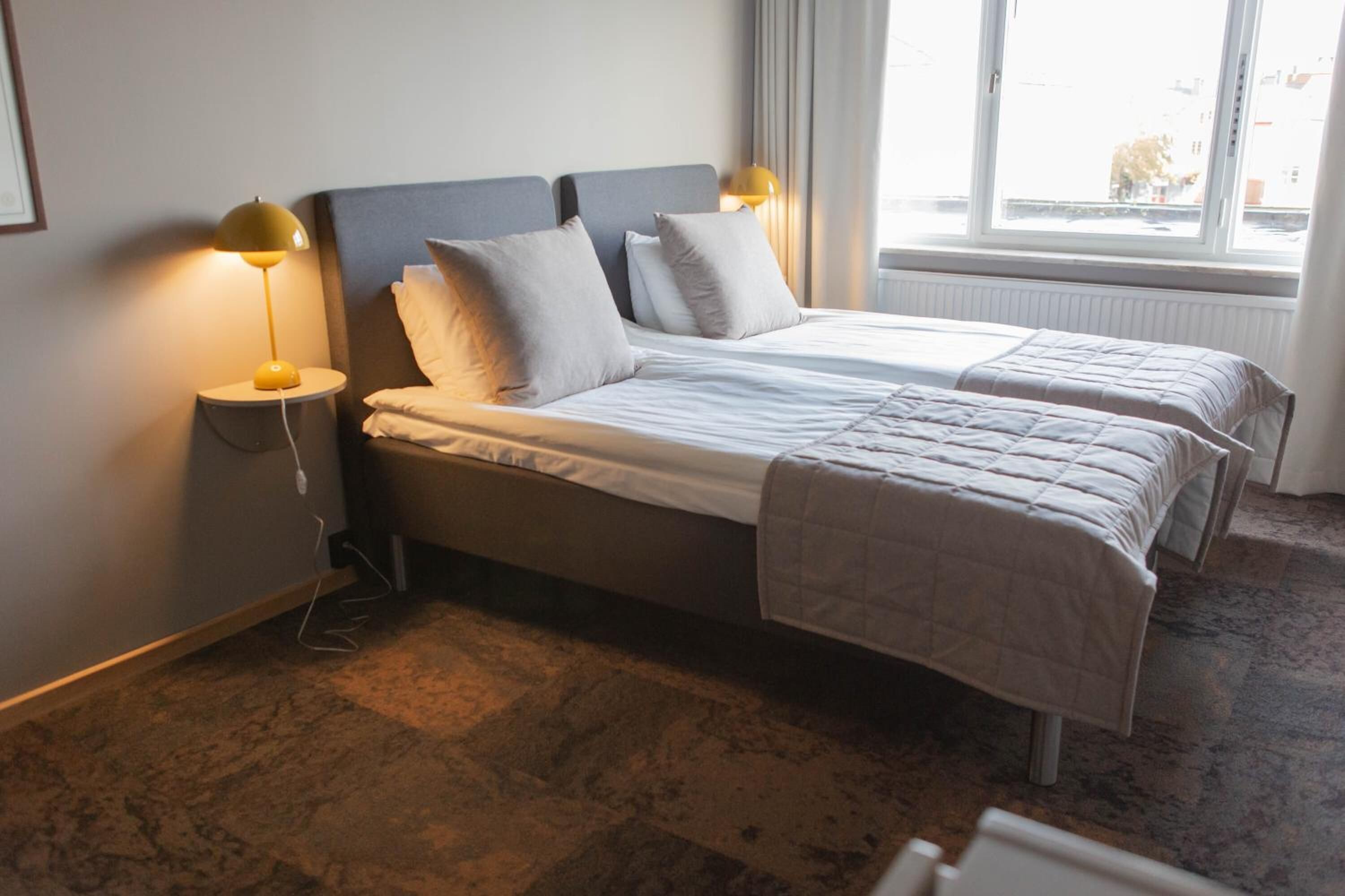Una cama doble con cabecera gris y ropa de cama blanca, dos lámparas de noche y una ventana con vistas al exterior.