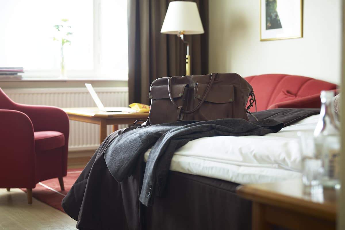 A hotel room with a bed, a red armchair, a desk with a laptop, a lamp, and a handbag on the bed.
