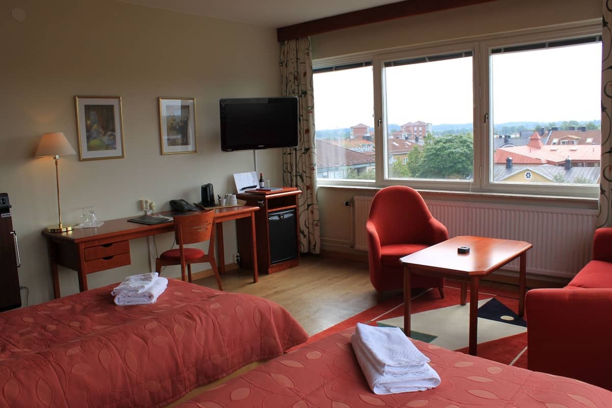 A hotel room with two beds, a desk, a chair, a TV, and a view of buildings through the window.