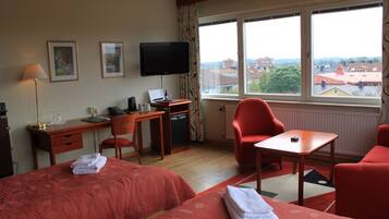 Habitación de hotel con dos camas, un escritorio, una silla, un televisor y vistas a edificios a través del ventanal.