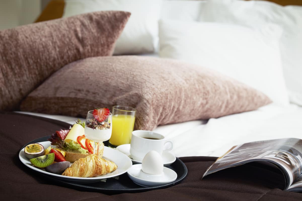 A neatly arranged breakfast tray on a bed with pillows, including croissants, fruits, yogurt, and a glass of orange juice.