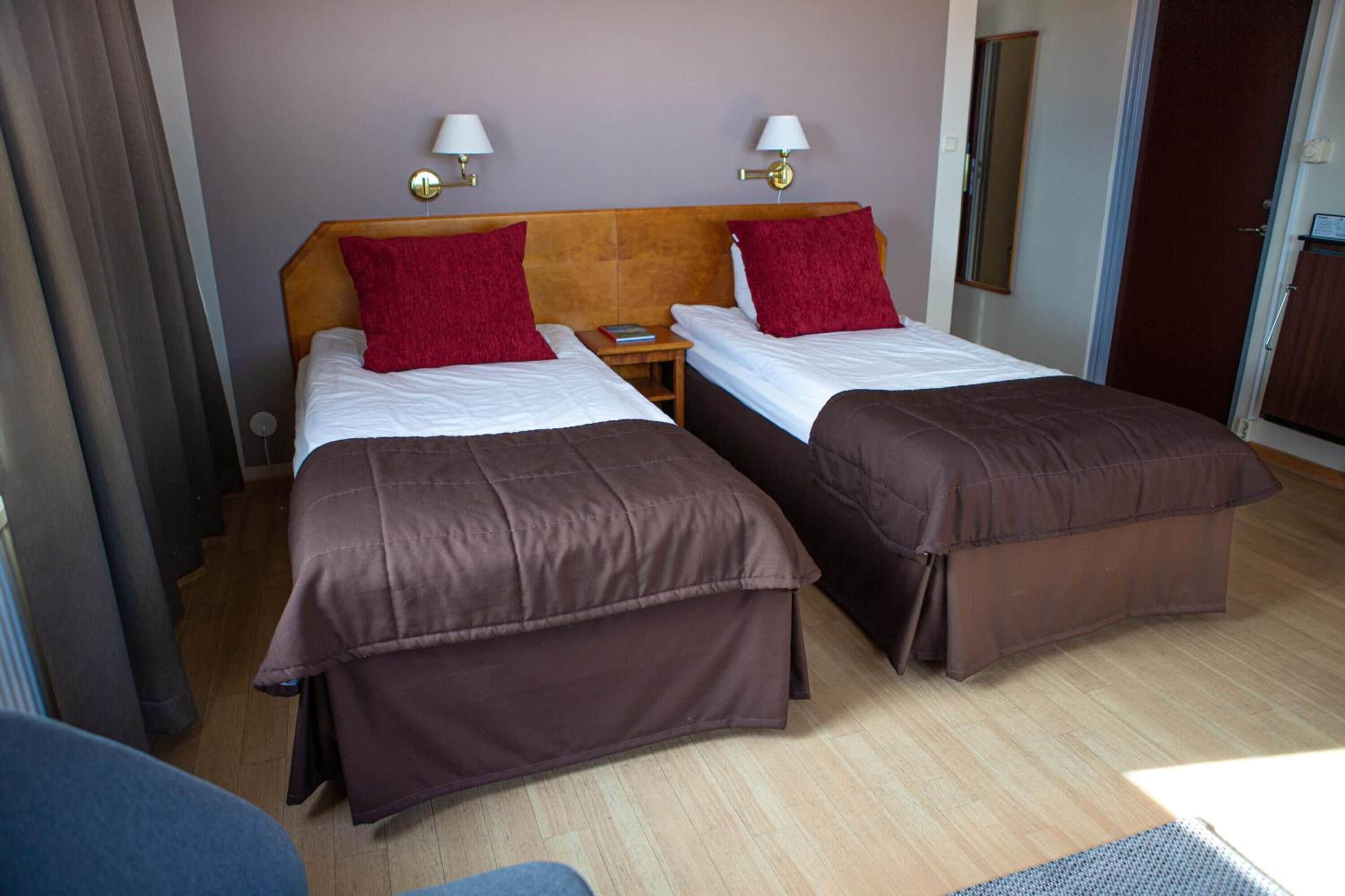Quarto de hotel com duas camas, cada uma com colcha marrom e travesseiro vermelho. Há mesinhas de cabeceira com luminárias e um criado-mudo pequeno.