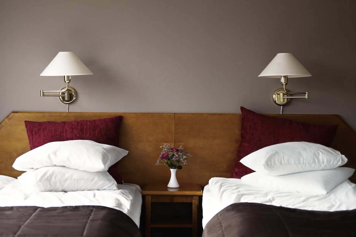 A hotel room with two beds, each with white linens, a wooden headboard, and red pillows. There are two wall-mounted lamps with white shades. A small white vase with flowers is placed between the beds.