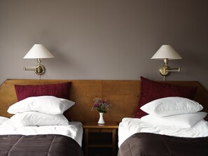 Habitación de hotel con dos camas, cada una con ropa de cama blanca, cabeceros de madera y almohadas rojas. Hay dos lámparas de pared con abatidores blancos. Un pequeño jarrón blanco con flores está colocado entre las camas.