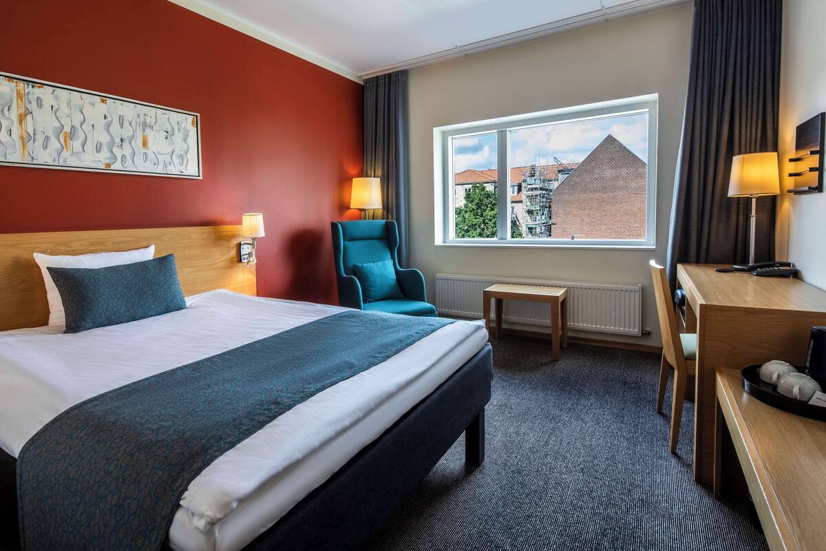 A hotel room with a large bed, a desk, a chair, a window with a view of buildings, and a painting on the wall.